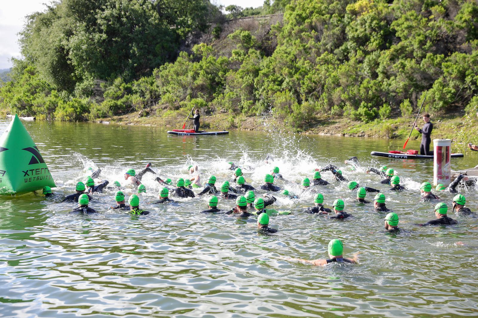 A group of swimmers wearing green swim caps compete in open water near a buoy. The lush, green landscape surrounds the lake, and two people on paddleboards are nearby, overseeing the event.