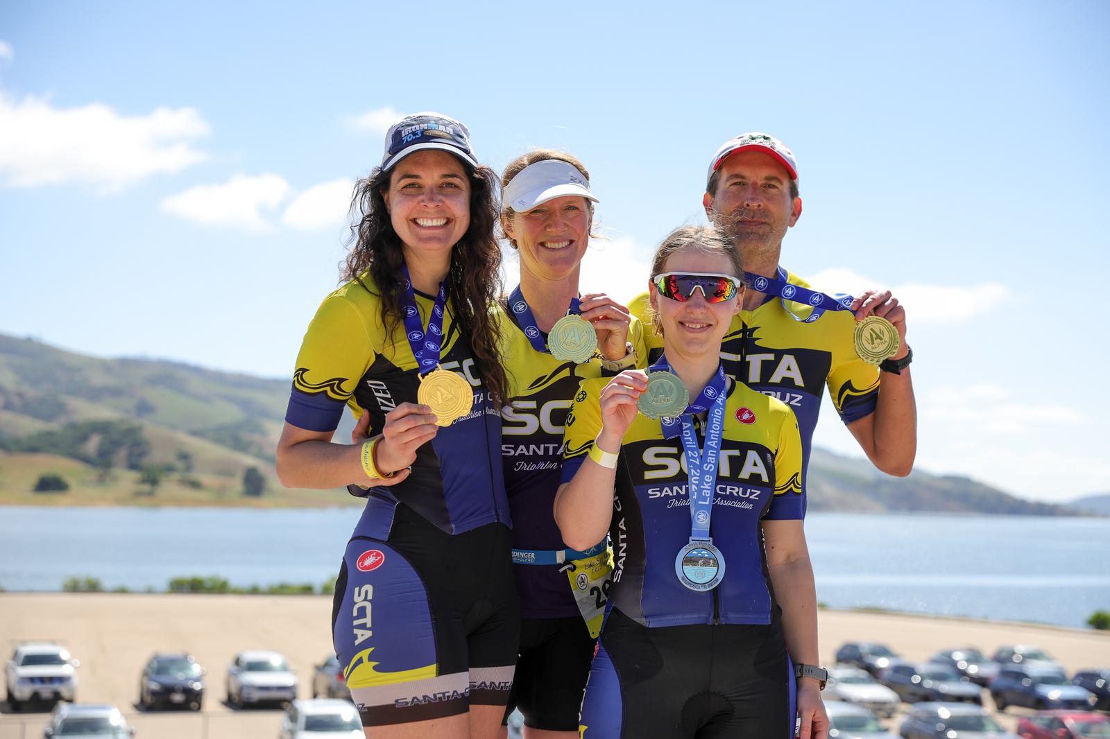 Four cyclists wearing matching "STA Santa Cruz" jerseys proudly display their medals. They are standing outdoors near a body of water, with rolling hills and parked cars in the background. The sky is clear and sunny.