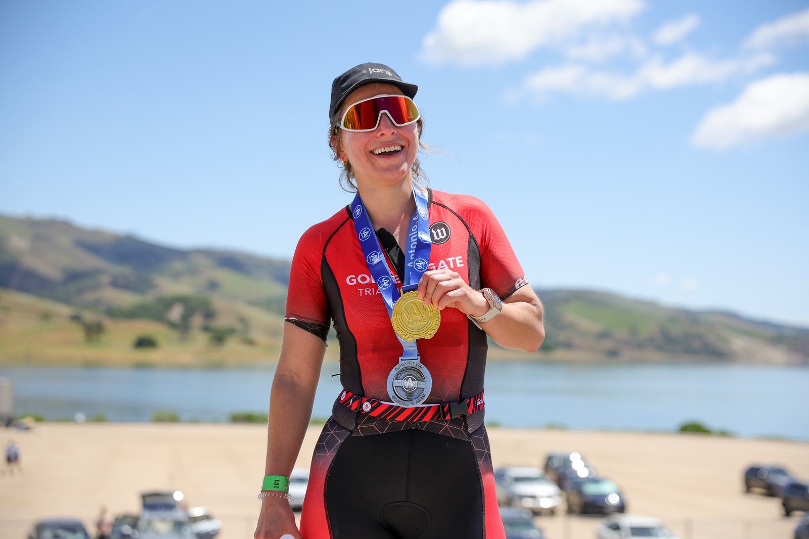 A cyclist in a red and black outfit stands outdoors, smiling and holding a gold medal around their neck. They are wearing sunglasses and a cap. The background features a scenic landscape with hills, a large body of water, and a blue sky with clouds.