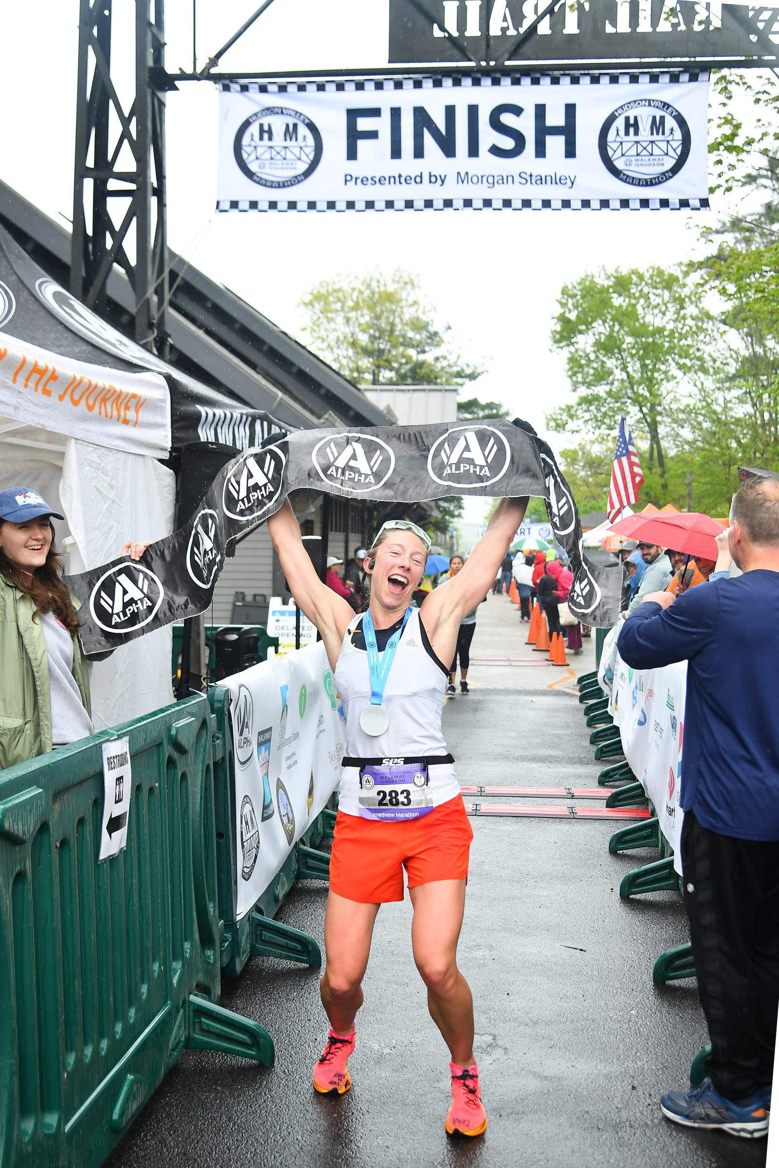A runner joyfully crosses the finish line, holding up a victory banner. They wear a white tank top, orange shorts, and a medal around their neck. Spectators cheer on either side under a gray sky.