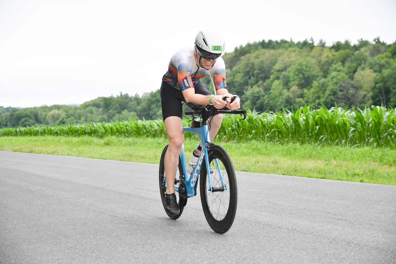 A cyclist wearing a white helmet and colorful jersey rides a blue bicycle on a paved road. Lush green foliage and trees are visible in the background. The setting is outdoors, likely during a race or training session.