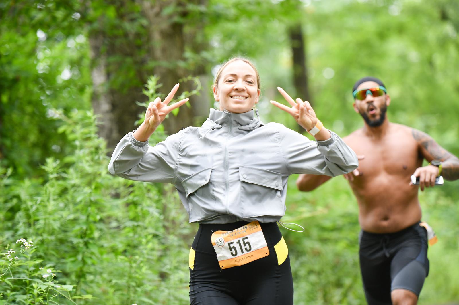 A woman smiling and gesturing with peace signs while running in a lush, green outdoor setting. She wears a gray jacket and has a race bib numbered 515. Behind her, a shirtless man in sunglasses runs along the same trail.