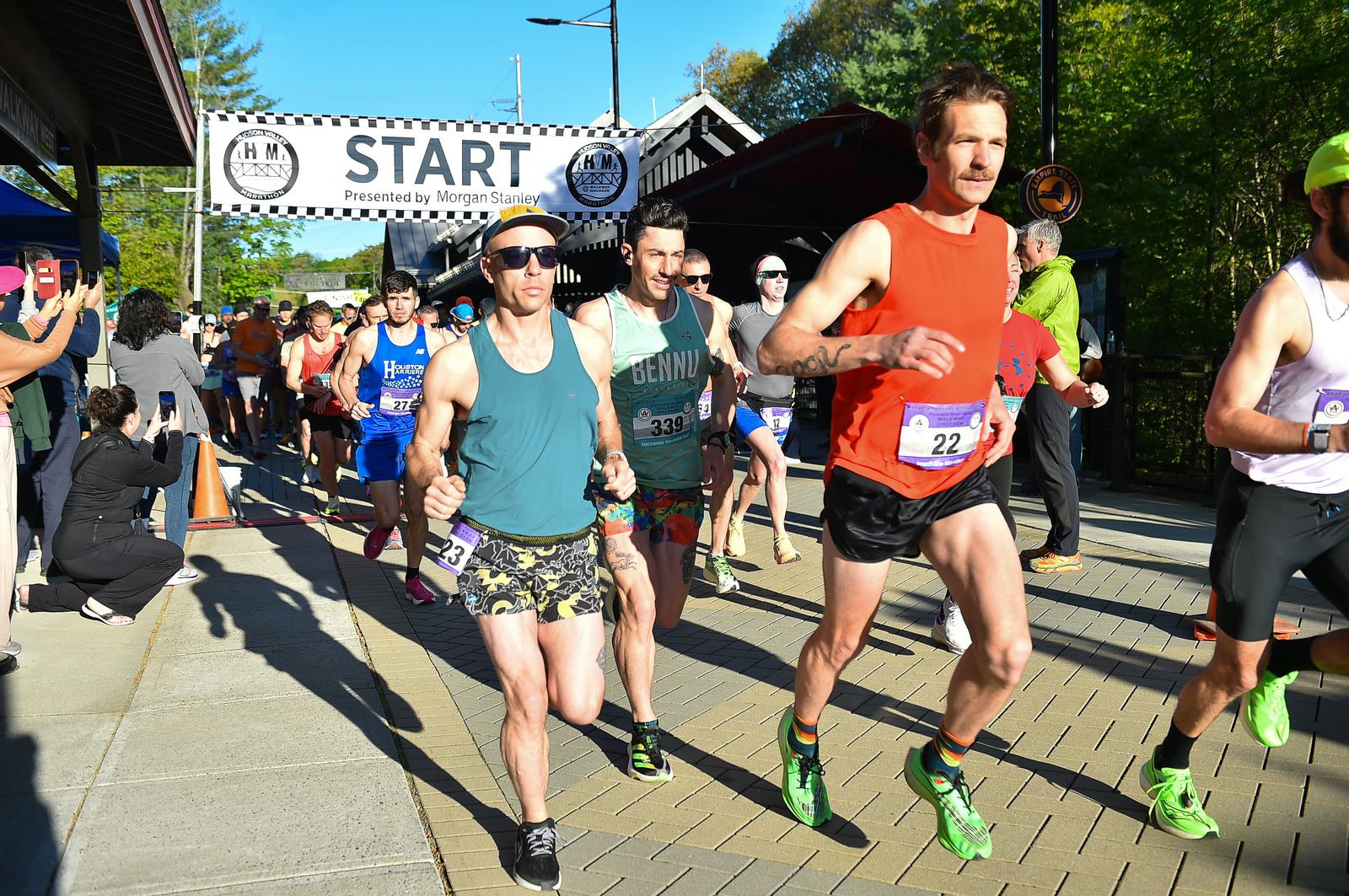 Runners in sporty attire take off at the starting line of an outdoor race on a sunny day. A large "START" banner is visible overhead, with spectators watching from the side. Trees and a pathway are in the background.