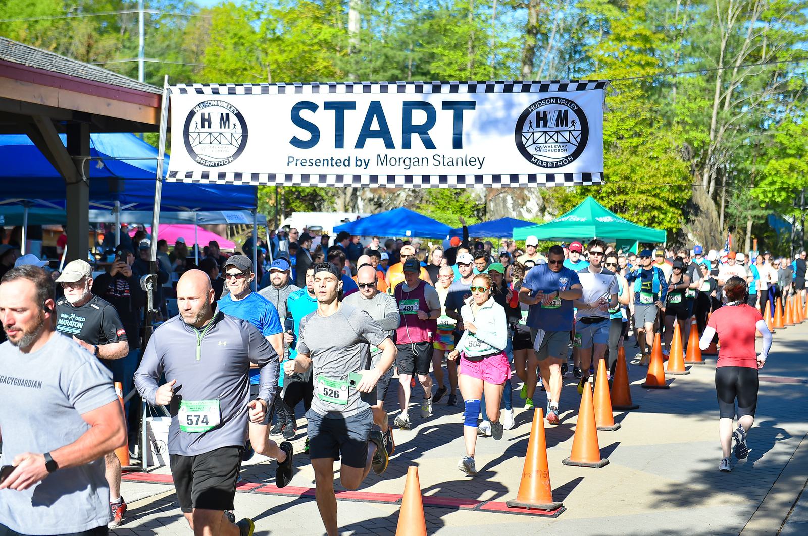 Runners in athletic gear begin a race under a "START" banner on a sunny day. The event is sponsored by Morgan Stanley. Orange cones line the pathway, and trees and colorful tents are in the background.