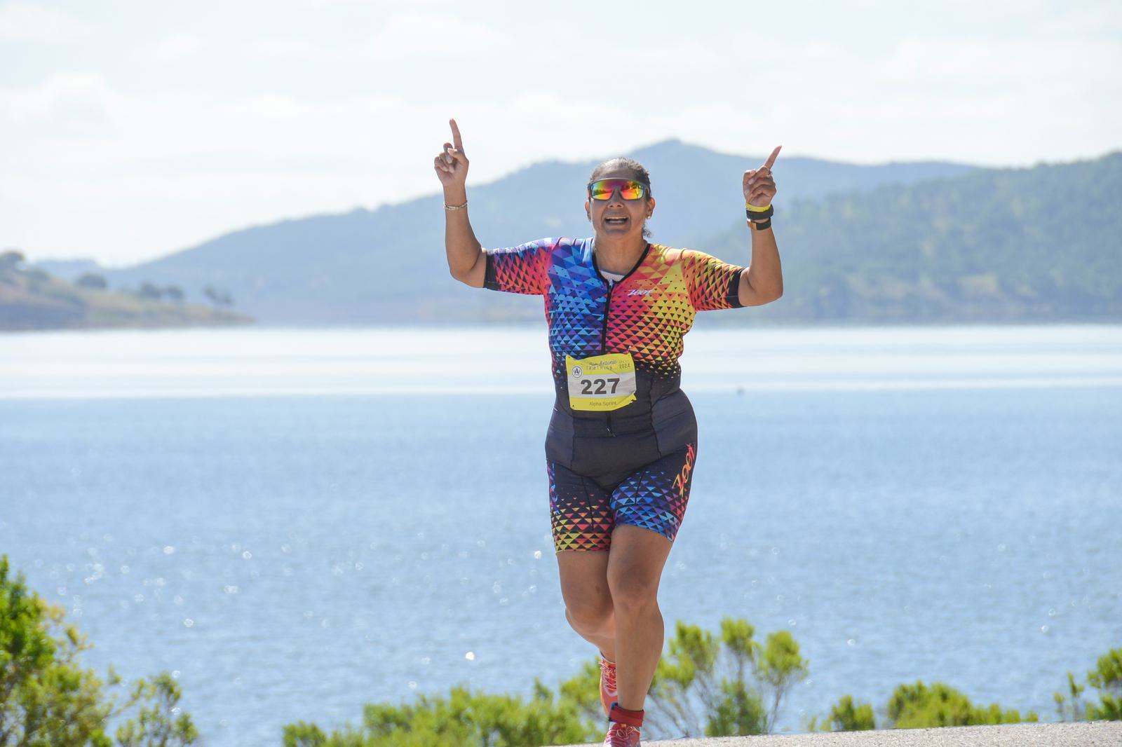 A runner wearing a colorful outfit and sunglasses smiles while jogging by a body of water. She raises both hands with pointed fingers. Hills and trees are visible in the background.