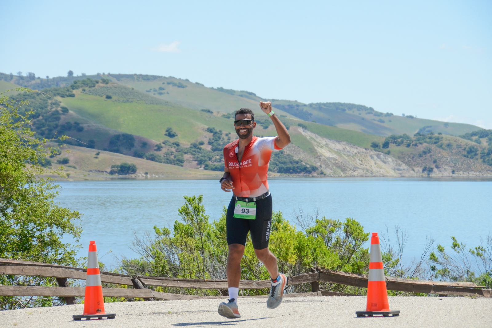 A man in a triathlon outfit runs on a path next to a lake, raising his fist in the air with a smile. Traffic cones line the path, and green hills are visible in the background under a clear blue sky.