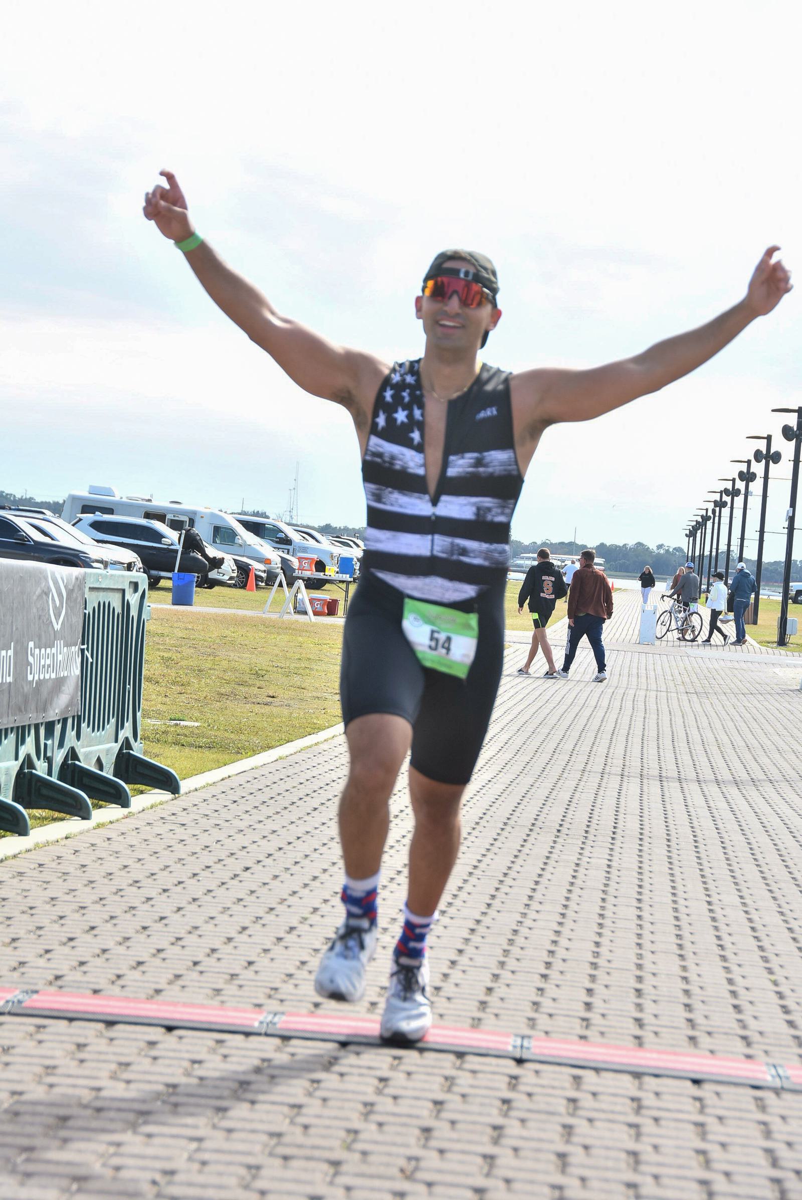 A person wearing a sleeveless outfit with a flag design crosses a finish line with arms raised. They are on a paved path, with a grassy area and parked cars in the background. A few other people are walking nearby.