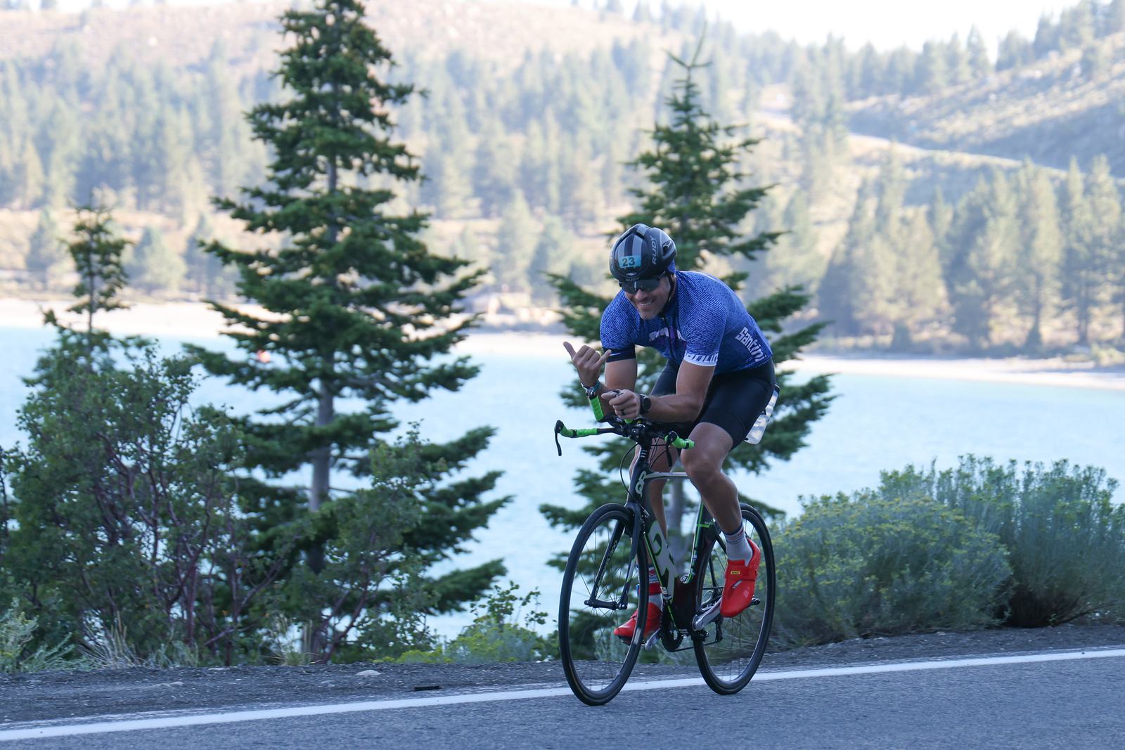 A cyclist wearing a blue jersey, black shorts, and helmet rides a bike on a road next to a scenic lake and pine trees. The background features hills and a clear sky.