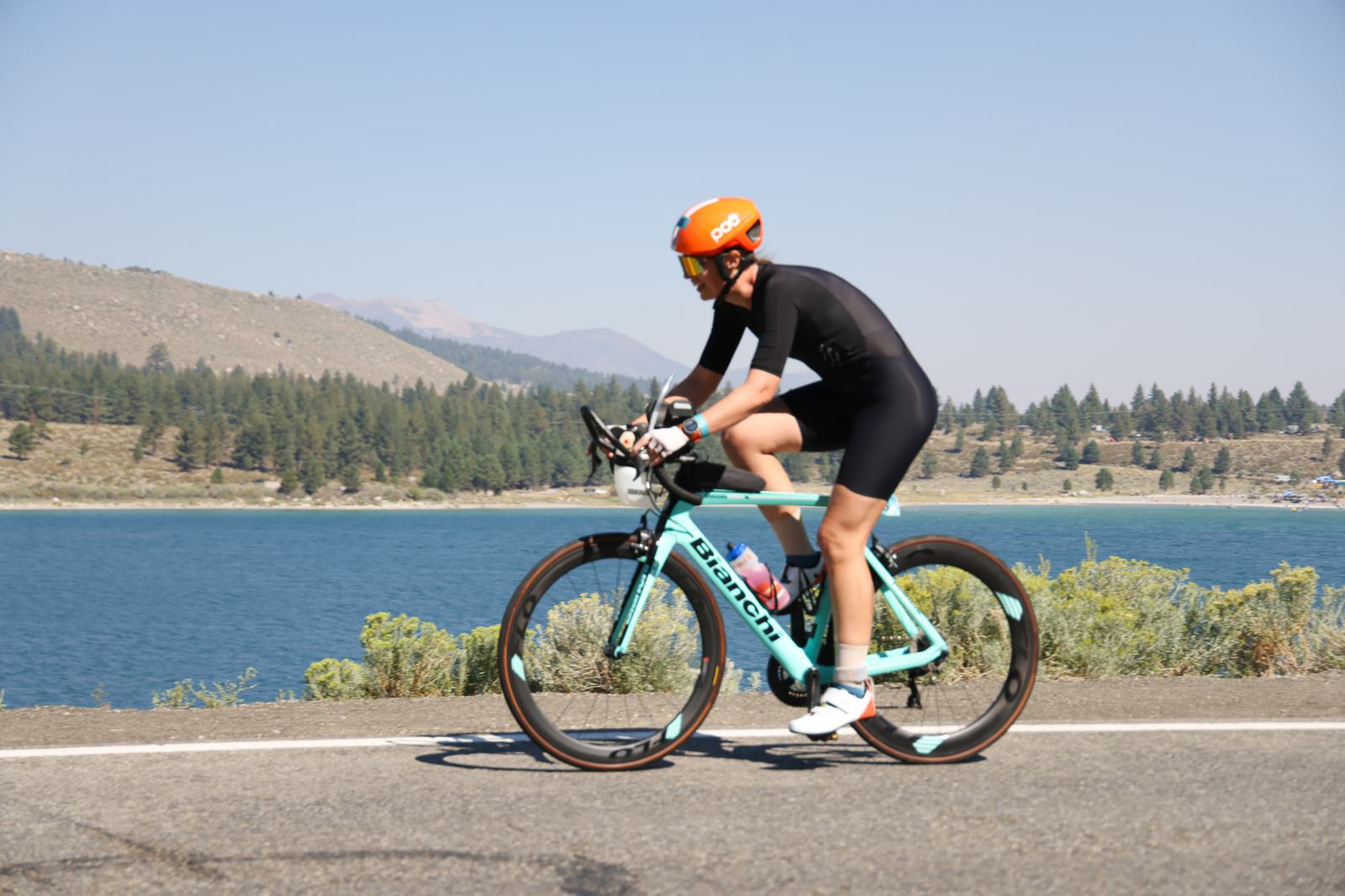 Cyclist in an orange helmet and black cycling suit rides a turquoise bike on a road beside a blue lake. Trees and mountains are visible in the background under a clear sky.