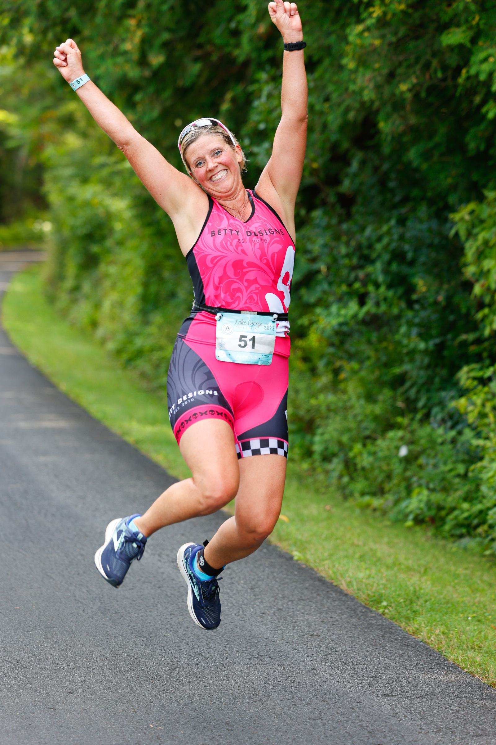 A woman wearing a pink and black athletic outfit jumps joyfully on a paved path. She has both arms raised, smiling widely, with a race bib numbered 51 on her outfit. Green trees line the path.