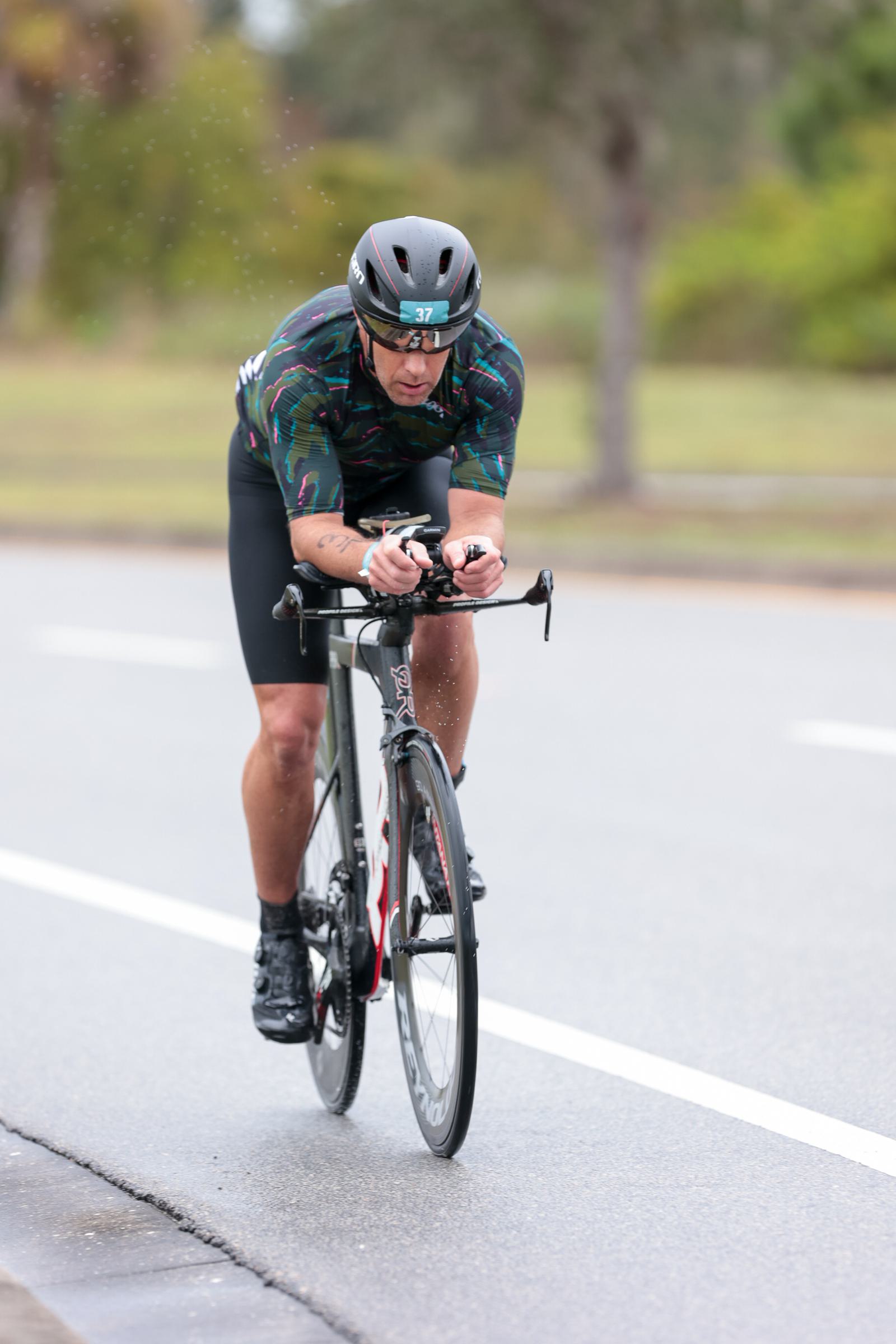A cyclist wearing a helmet and patterned jersey rides a racing bicycle on a wet road. The background is blurred, indicating motion.