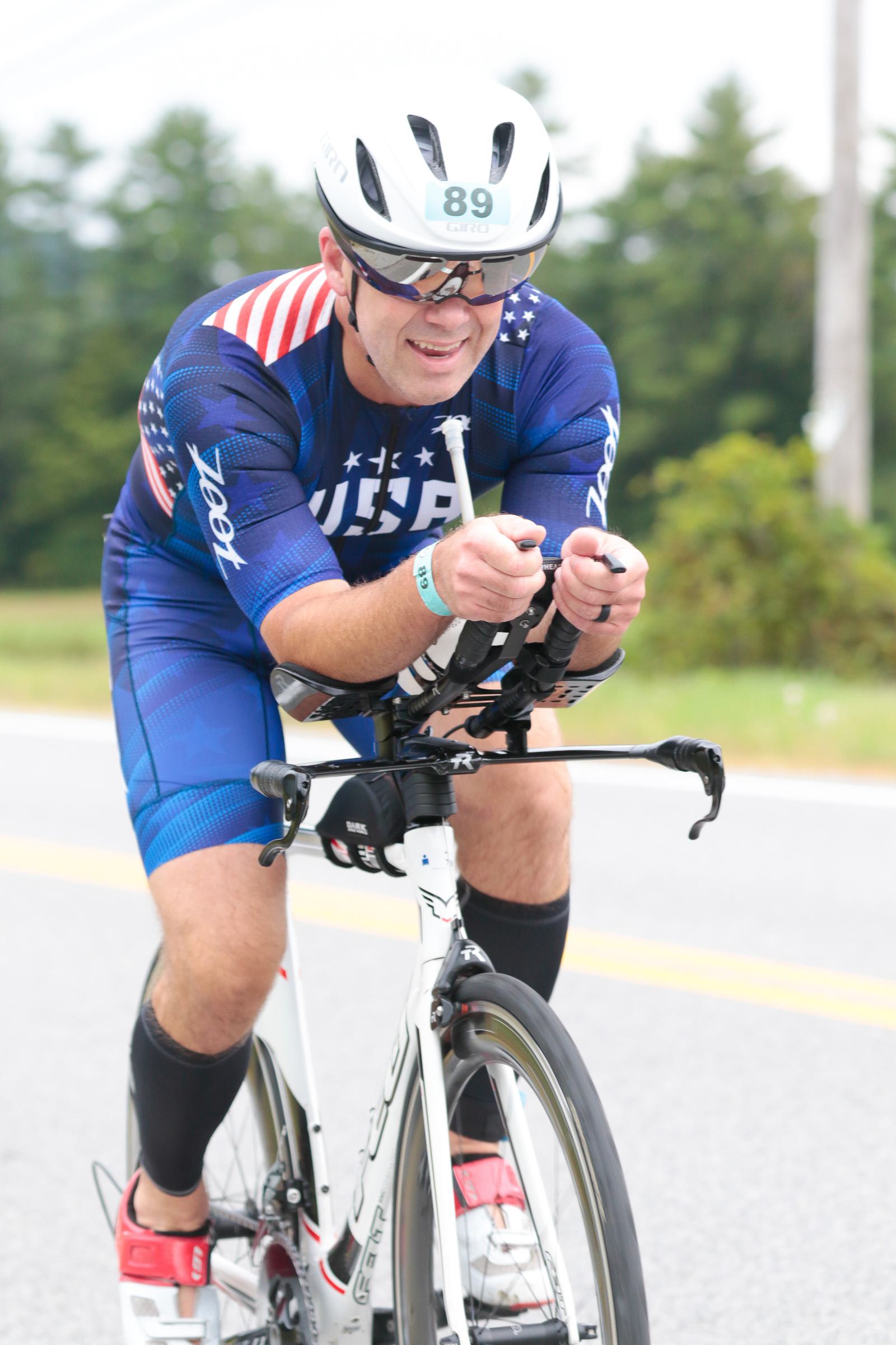 Cyclist wearing a USA-themed kit and helmet in a race, riding a white road bike on a paved road. The cyclist is focused, arms resting on aero bars, with a background of trees and cloudy sky.