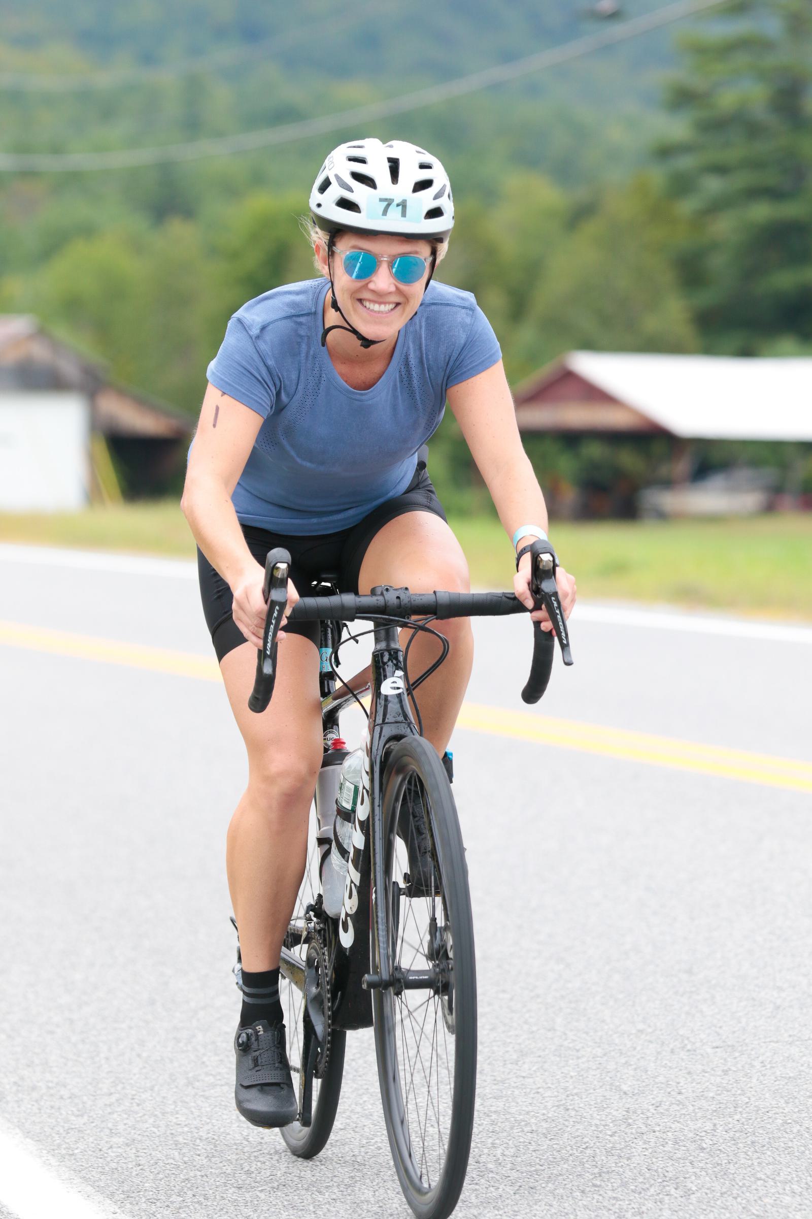 A cyclist in a blue shirt and helmet rides along a scenic road. The person is smiling, wearing sunglasses, and seems to be enjoying the ride. The background includes blurred green trees and hills.