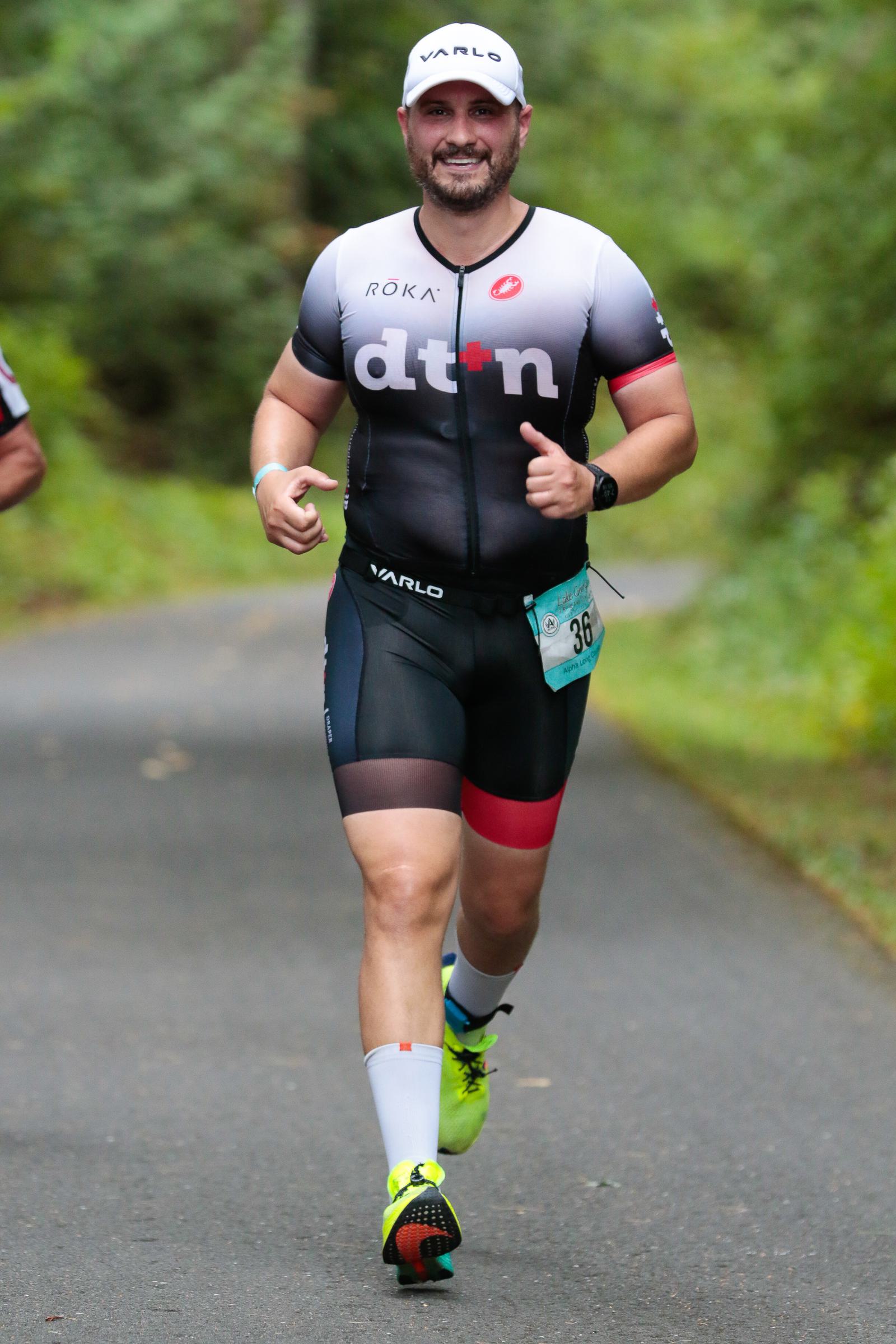 A person is running on a paved path surrounded by greenery, wearing a black and gray athletic outfit with red accents, a white cap, and bright yellow sneakers. The runner has a race bib pinned to the shorts.