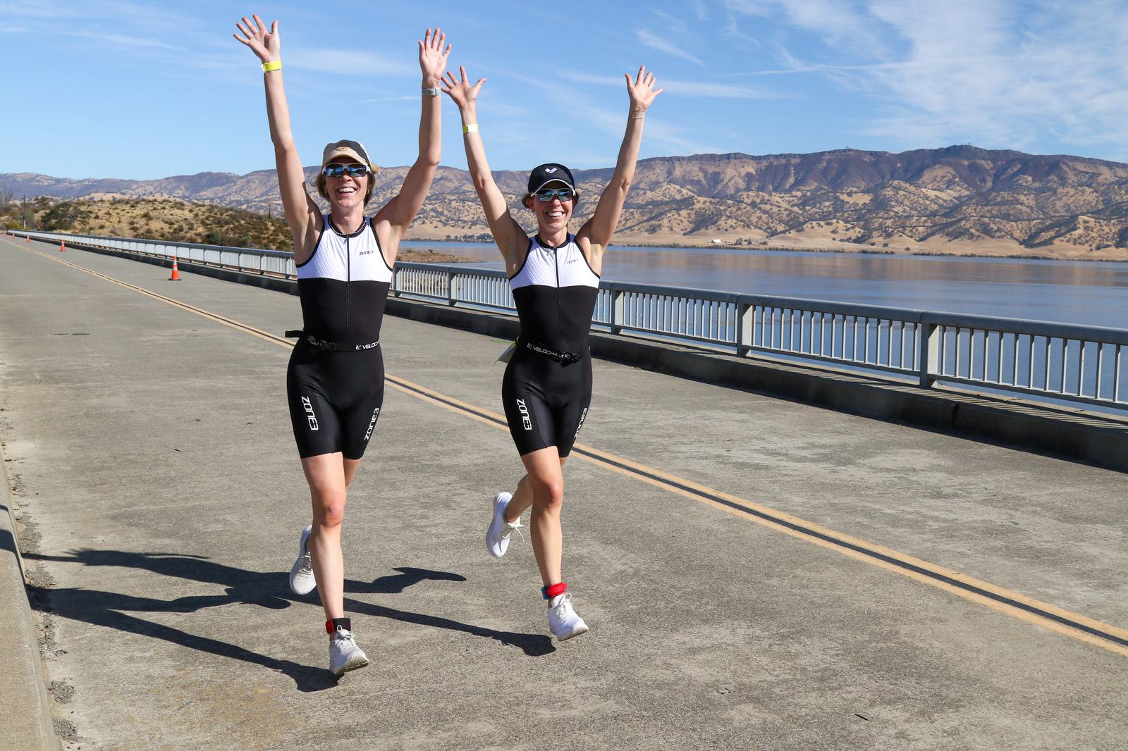 Two athletes in matching triathlon outfits and sunglasses joyfully raise their arms while running along a scenic lakeside road. In the background, hills and a clear blue sky add to the picturesque setting.