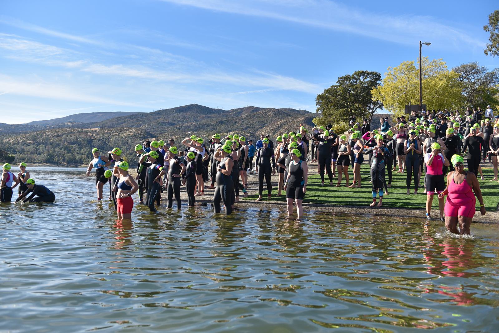 A large group of people in wetsuits and green swim caps gather by a lake, preparing for a swim. Some are entering the water while others wait on the grassy shore. Trees and hills are in the background under a clear blue sky.