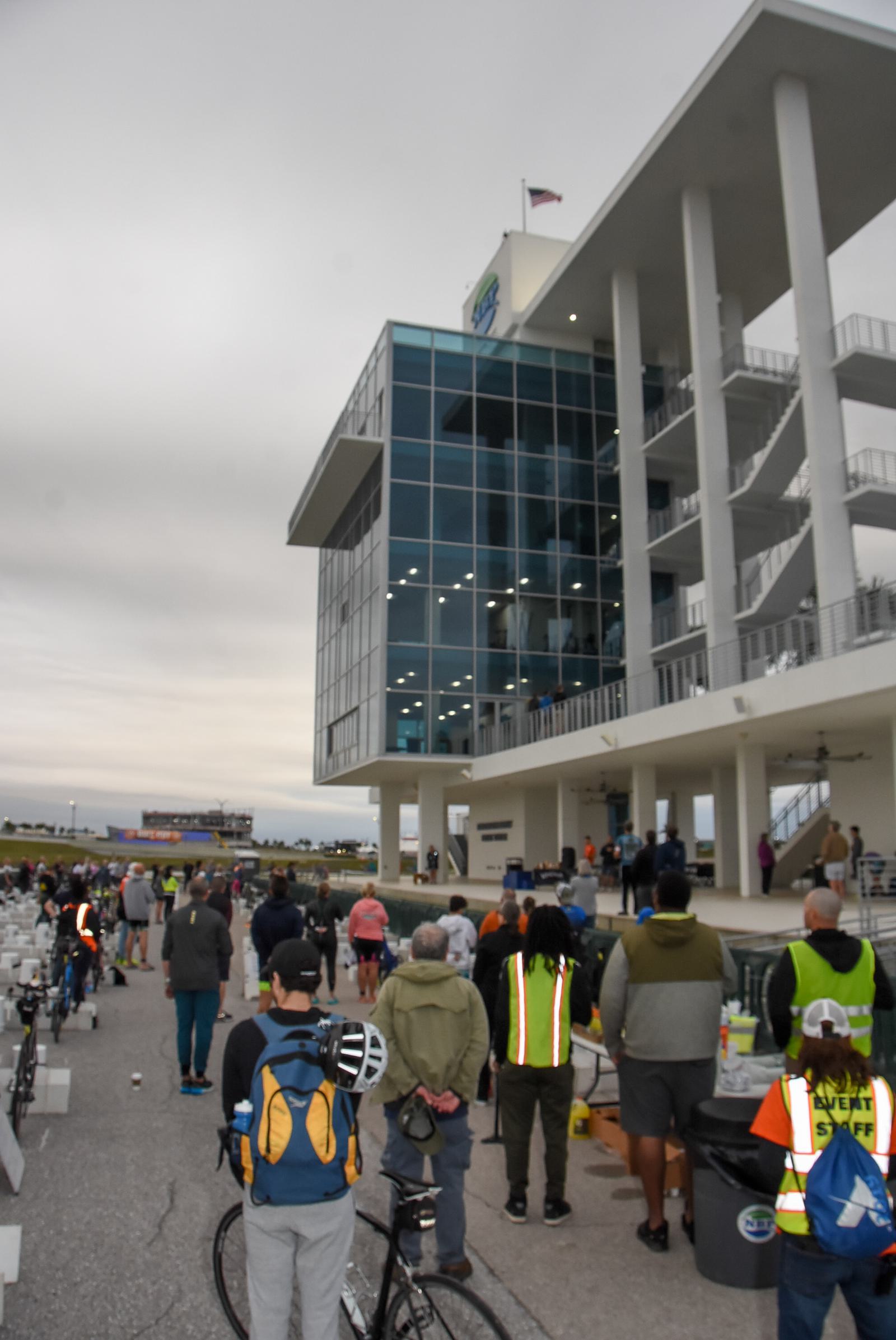 A group of people standing outside a modern building with a glass facade and tall pillars. Some hold bicycles, while others wear reflective vests. The sky is overcast, and a flag flies atop the building.
