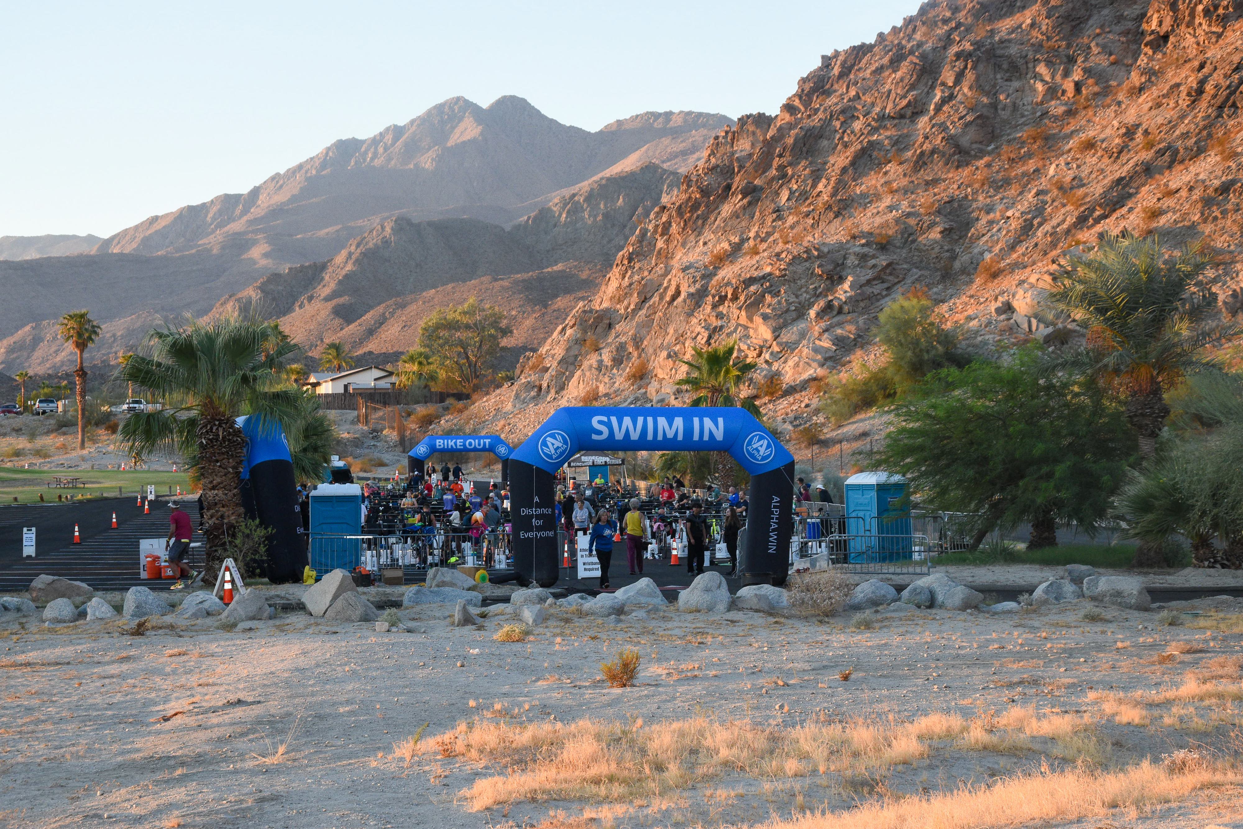 A group of people gathers at the "Swim In" transition zone during a triathlon, set against a backdrop of rocky mountains and desert landscape. The area is marked by a large inflatable archway, with palm trees and dry terrain surrounding it.