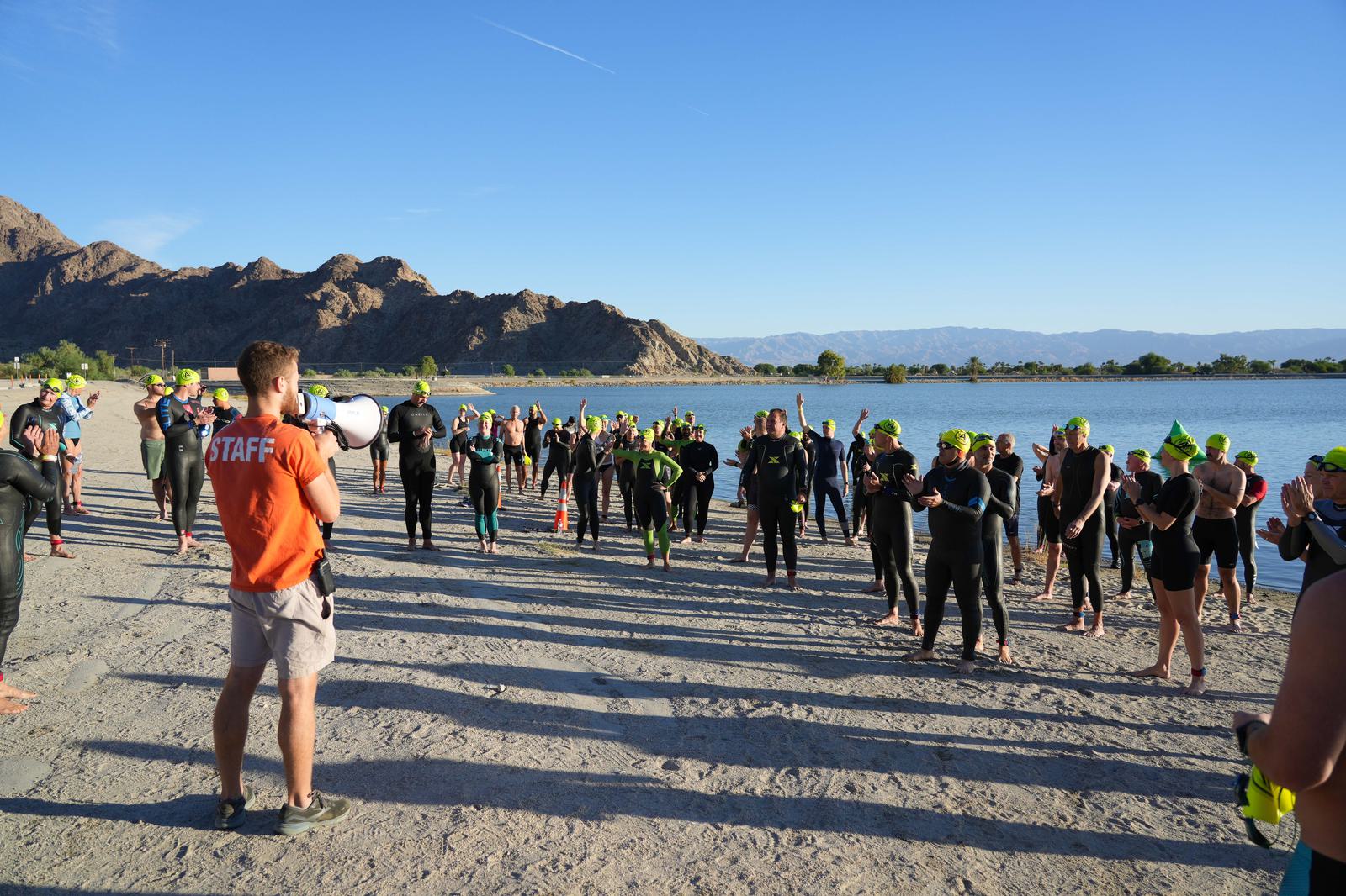 A group of people in wetsuits and green swim caps stand on a sandy beach, listening to a person in an orange shirt with a megaphone. Rocky hills and blue sky are in the background.