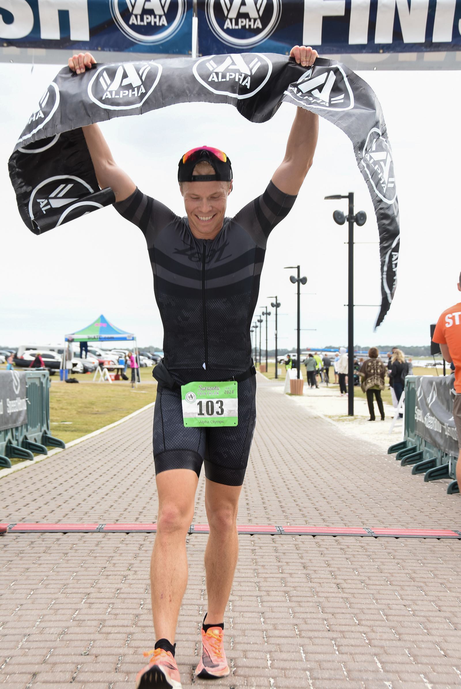 Athlete in a black outfit joyfully crosses the finish line, holding a banner. They're on a paved path with a green race bib numbered 103. Spectators and event tents are visible in the background.