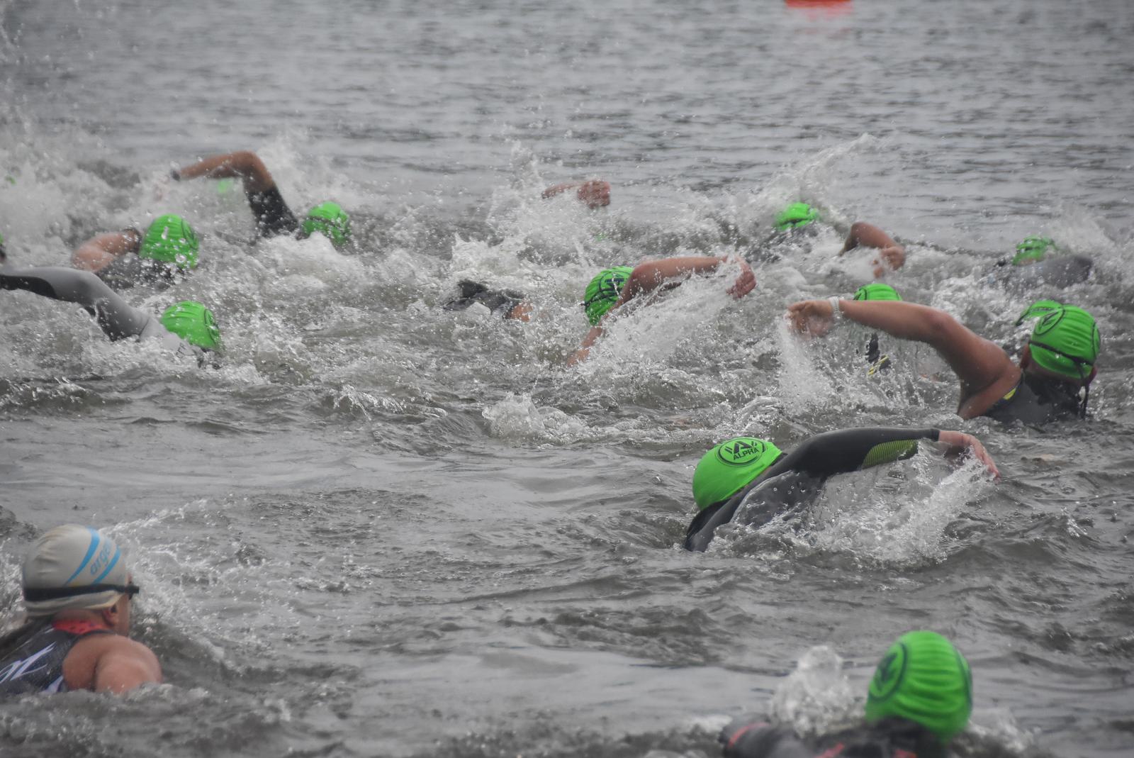 A group of swimmers in wetsuits and bright green swim caps competes in open water. The swimmers are mid-stroke, creating splashes as they move through the water. One swimmer wears a white cap. The scene conveys energy and competition.
