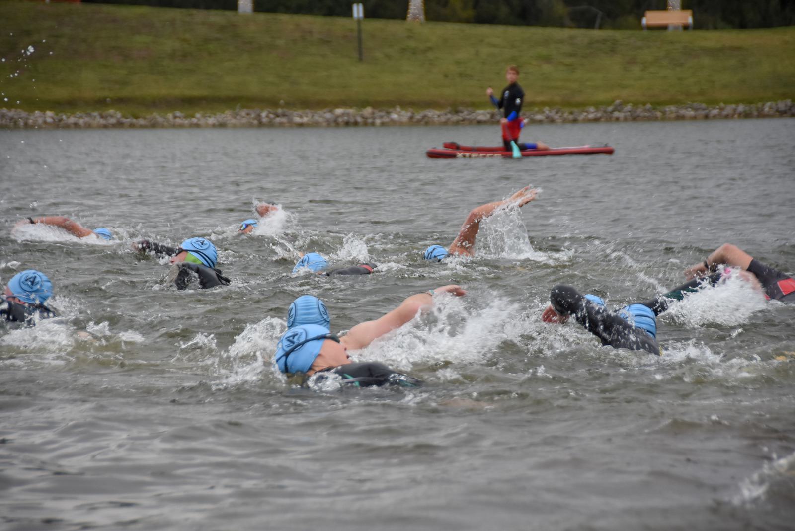 Swimmers in blue caps participate in an open water race, creating splashes as they stroke through the water. A person on a paddleboard observes them from a distance against a backdrop of grassy land.