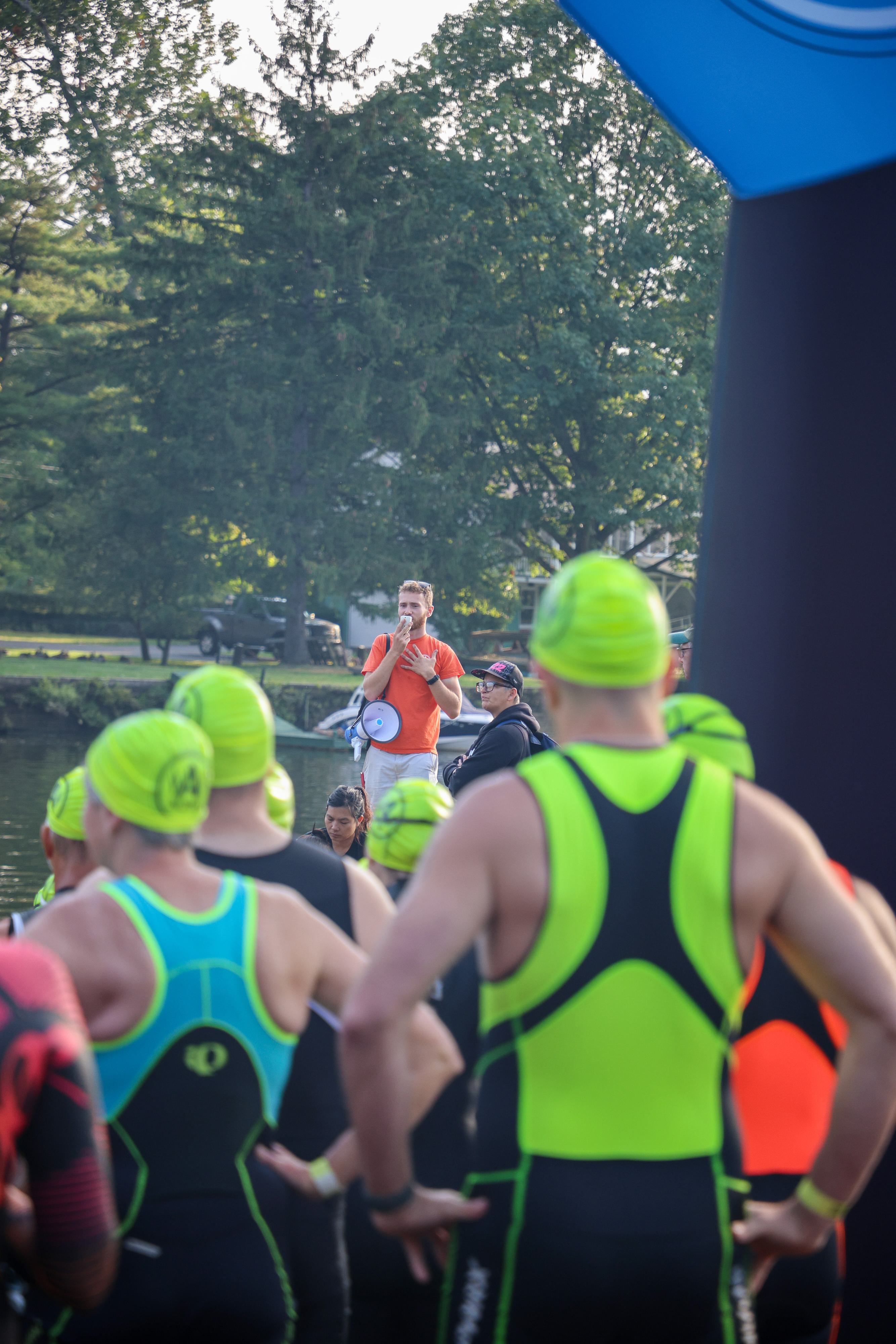 A group of athletes in bright green swim caps and wetsuits listen to a person with a megaphone near a lake, surrounded by trees. The speaker is on a platform, and it appears to be the start of an outdoor water event.