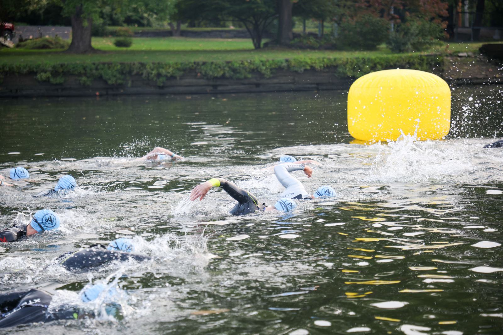 A group of swimmers wearing swim caps and wetsuits compete in open water. They swim toward a large yellow buoy in a calm outdoor setting, surrounded by greenery. Splashes of water indicate their swift movement.