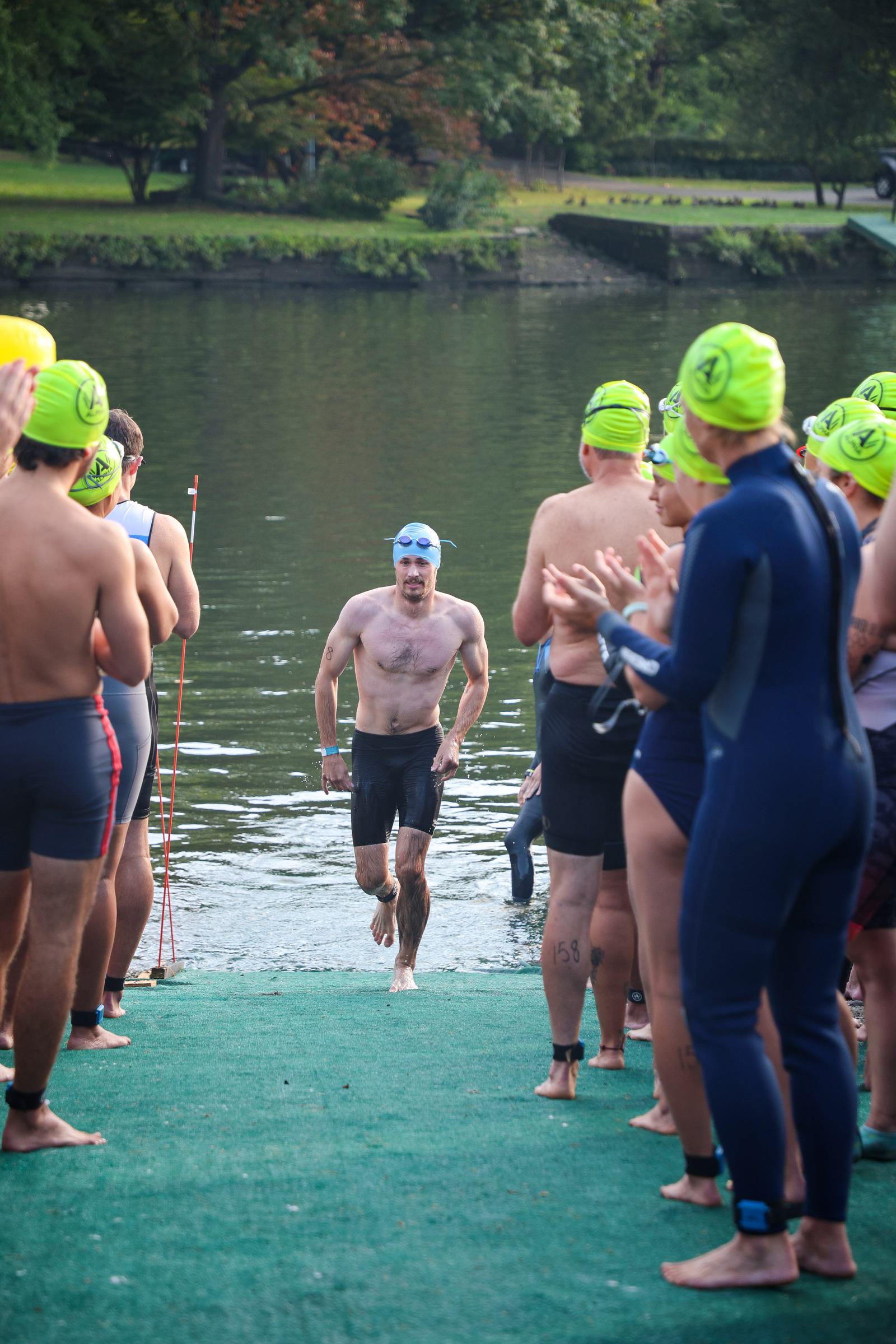 A male triathlete in a blue swim cap emerges from a river, running between two lines of cheering people. They are wearing wetsuits and yellow or green swim caps. Trees and a riverbank are in the background.
