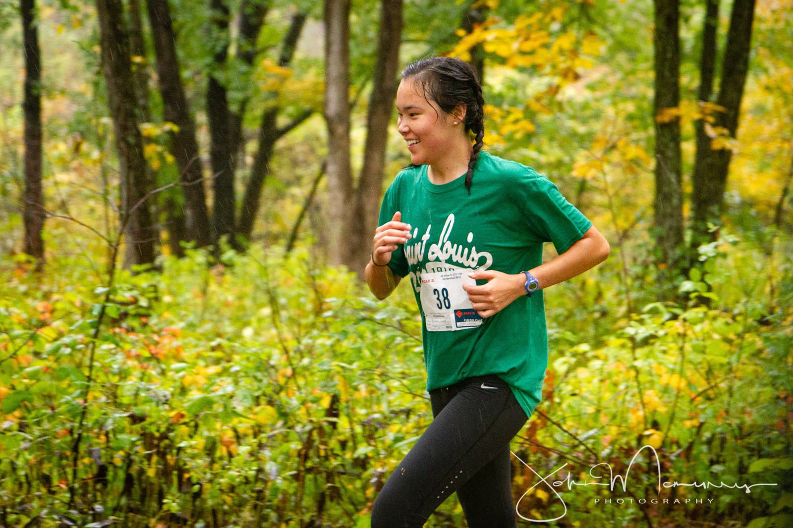 A person in a green shirt and black leggings jogs through a lush, green forested area. They are wearing a numbered race bib and have a cheerful expression. The tall trees and dense foliage in the background suggest a trail run or outdoor event.