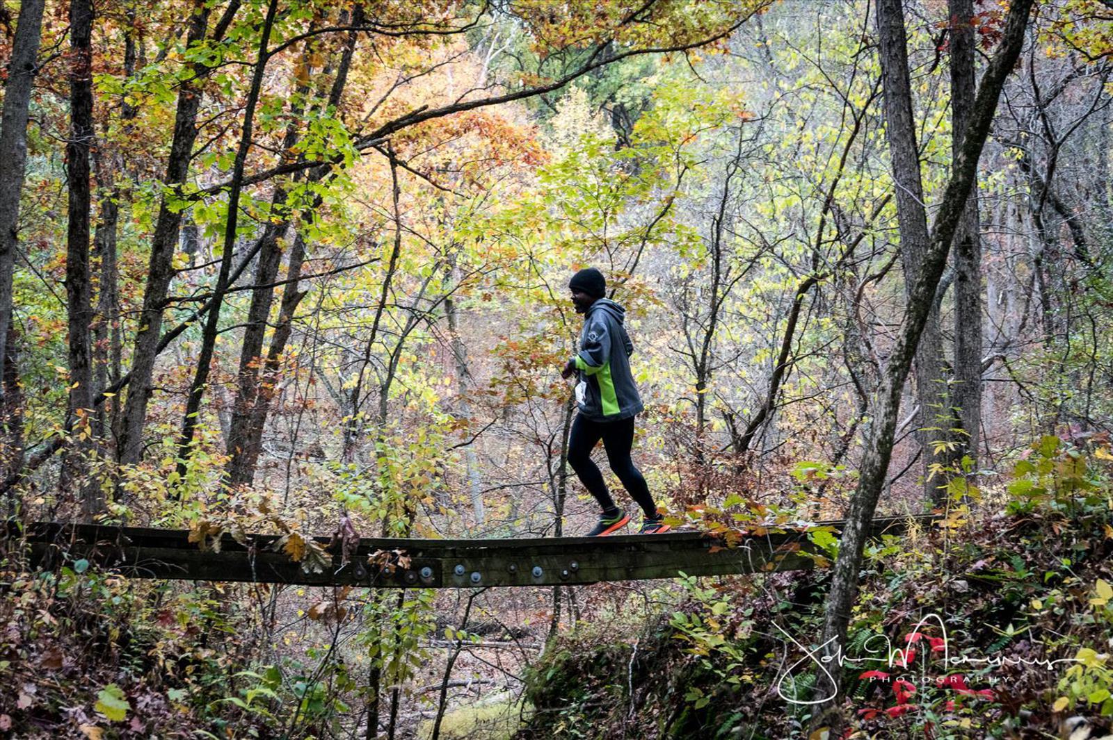 A person jogs on a narrow wooden bridge surrounded by a colorful autumn forest with leaves in shades of orange, yellow, and green. The person wears a dark outfit and sneakers. Trees and fallen leaves cover the ground around the serene path.