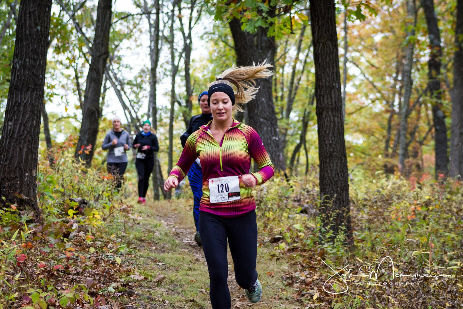 A woman wearing a colorful long-sleeve shirt and dark leggings runs through a forest trail, smiling. Other runners follow her in the background. The ground is covered with fallen leaves, and the trees have autumn foliage.