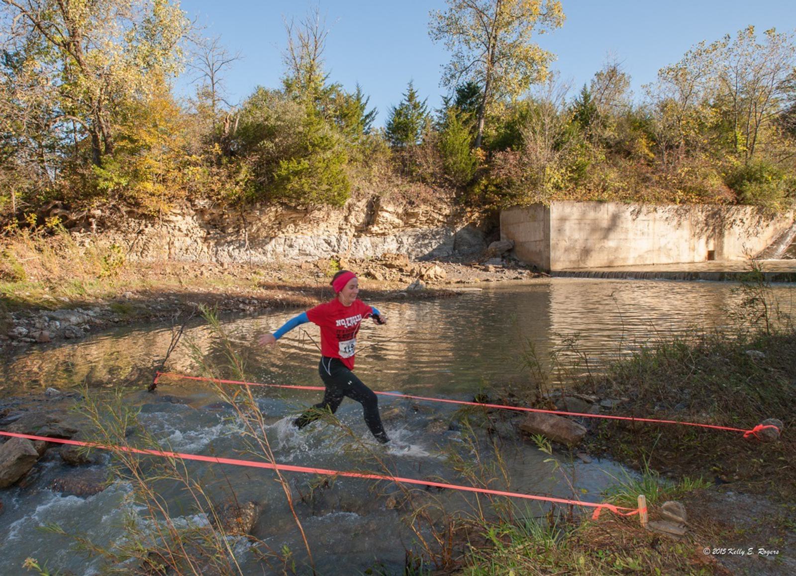A person in a red shirt and black pants runs energetically across a shallow stream, splashing water. They are surrounded by ropes and autumn trees, under a blue sky.