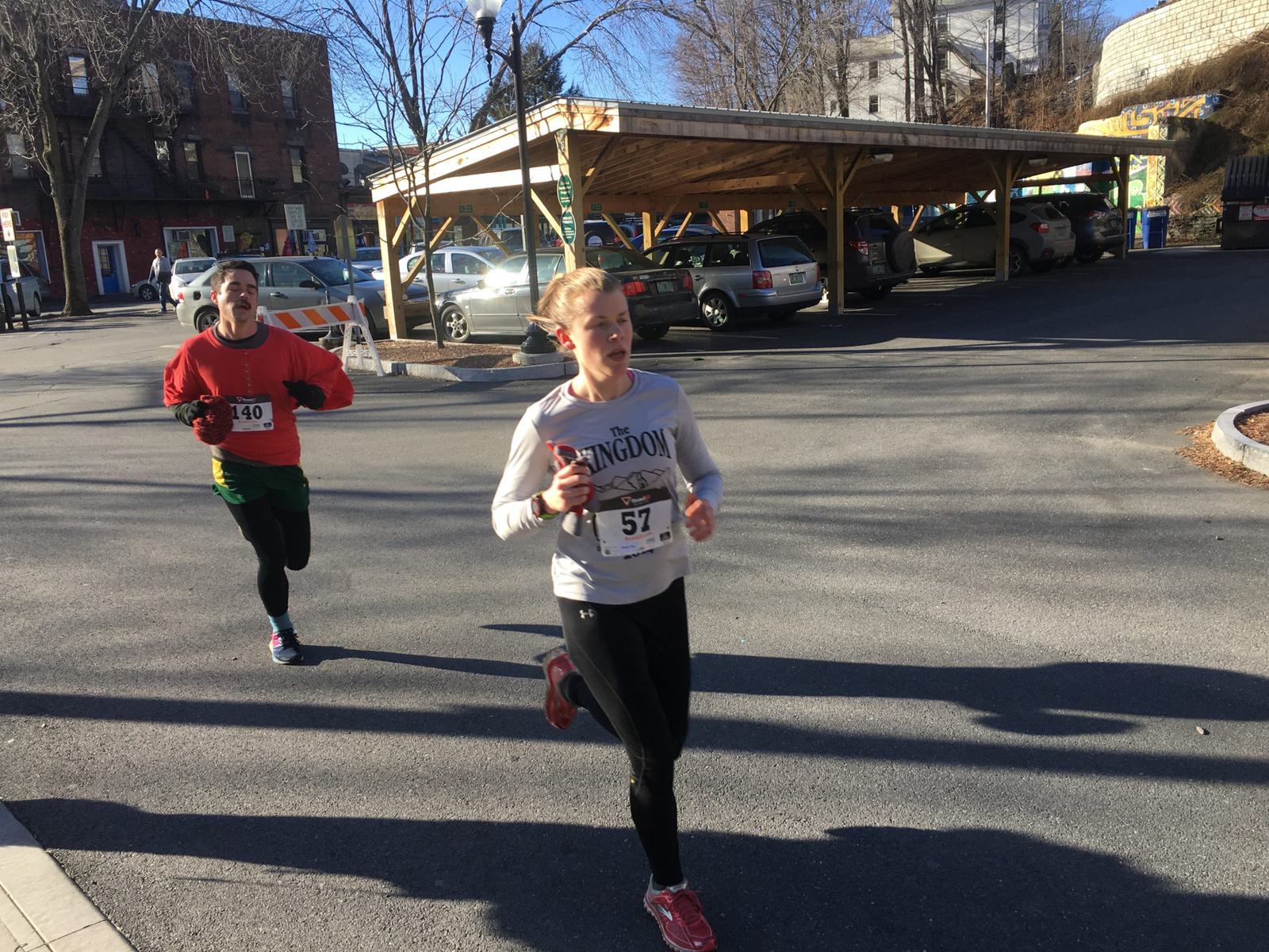 Two runners participate in a race on a sunny day. The woman in front wears a gray shirt and black leggings, while the person behind wears a red shirt. They are running on a paved road next to a parking area. Trees and buildings are in the background.