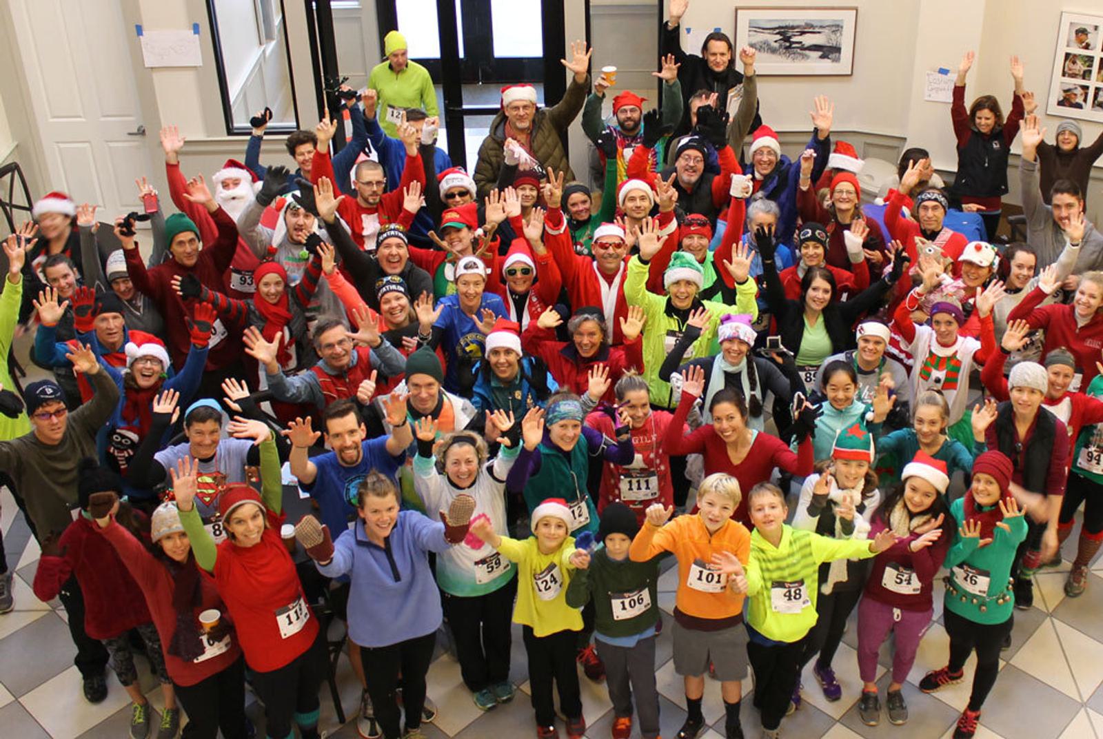 A large, cheerful group of people in colorful athletic gear, some wearing Santa hats, gather indoors for a holiday-themed race. They are waving and smiling at the camera, with race numbers visible on their outfits.