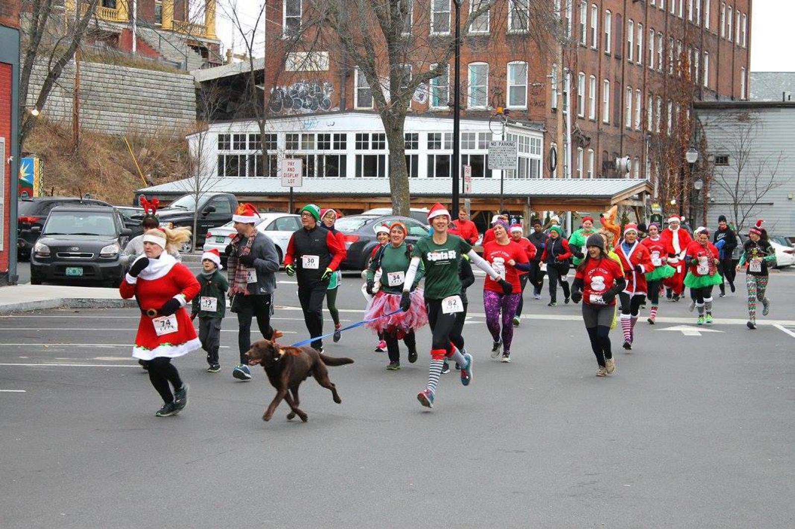 A group of people dressed in festive costumes, including Santa outfits and elf hats, participate in a holiday-themed fun run. They are jogging on a street, with some wearing race bibs and one person walking a dog on a leash. Buildings line the background.