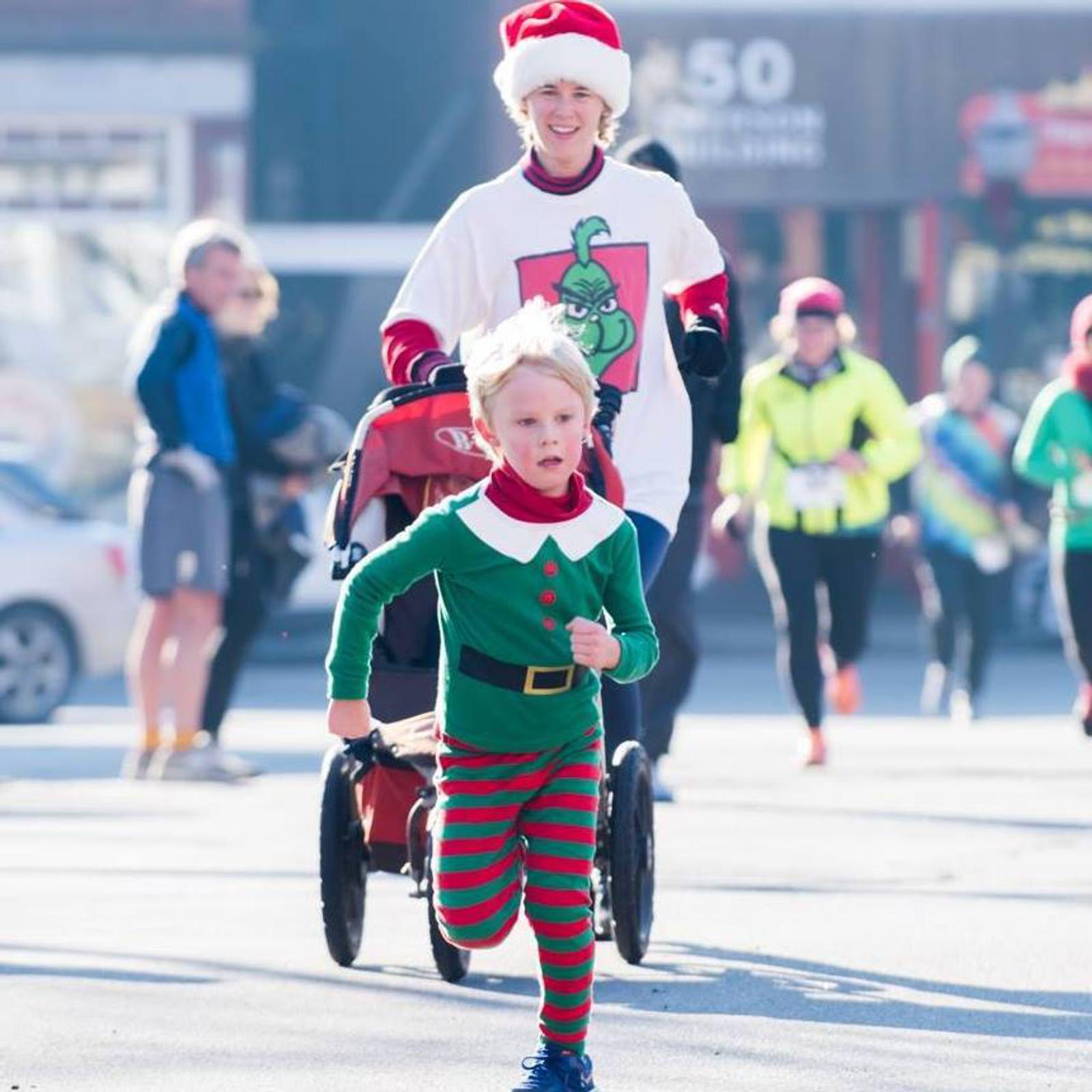 A child in a festive elf costume runs energetically in front of an adult wearing a Santa hat and pushing a stroller during a holiday-themed outdoor event. Other participants in festive attire are in the background.
