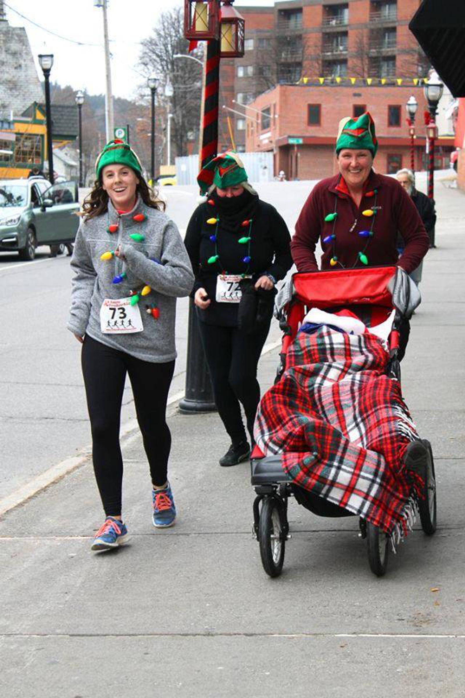 Three people in festive holiday attire jog on a city sidewalk. Two women jog side by side, while a third pushes a stroller covered with a red plaid blanket. Each wears an elf hat adorned with colorful lights. Cars and buildings are in the background.