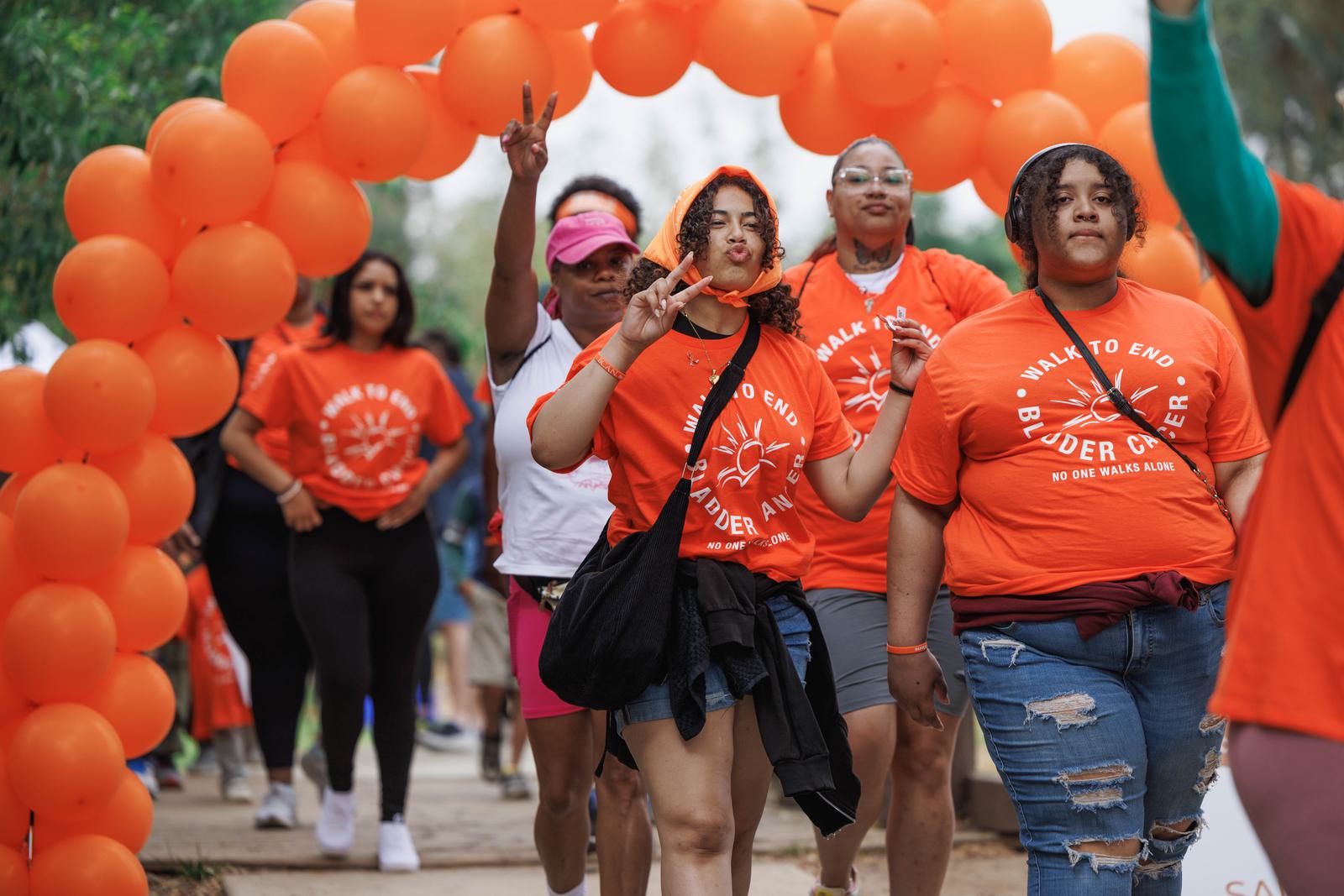 A group of people walks under an orange balloon arch, wearing matching orange T-shirts with a message about ending hunger. Some are making peace signs, and the mood is lively and positive.