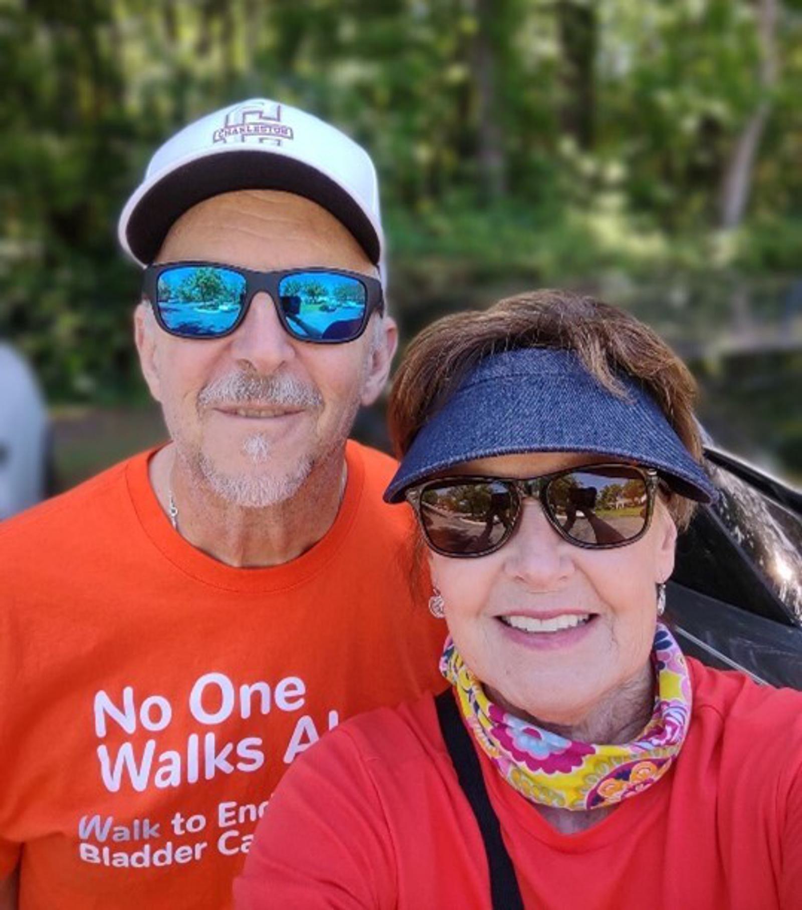 A man and woman pose outdoors with trees in the background. The man wears sunglasses, a white hat, and an orange shirt with text supporting bladder cancer awareness. The woman wears sunglasses, a visor, and a colorful scarf.
