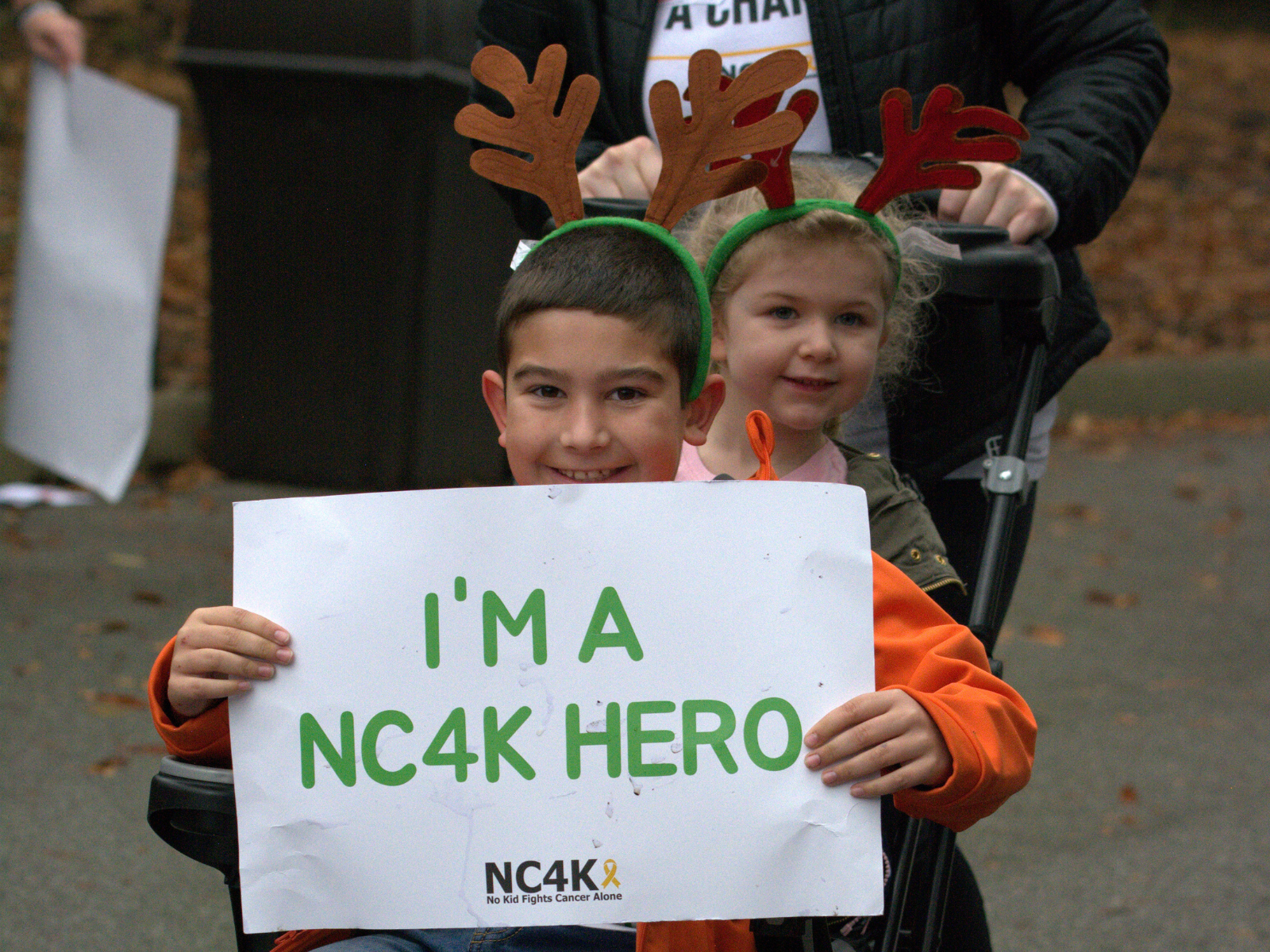 A young boy and girl wearing reindeer antlers hold a sign reading "I'm a NC4K Hero." They are seated in a stroller during an outdoor event.