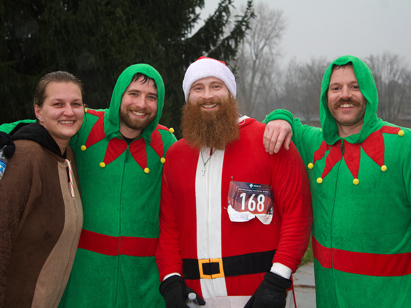 Four people in festive holiday costumes smiling outside. One is in a Santa suit, and the others are dressed as elves. The setting is overcast and appears to be outdoors with trees in the background.