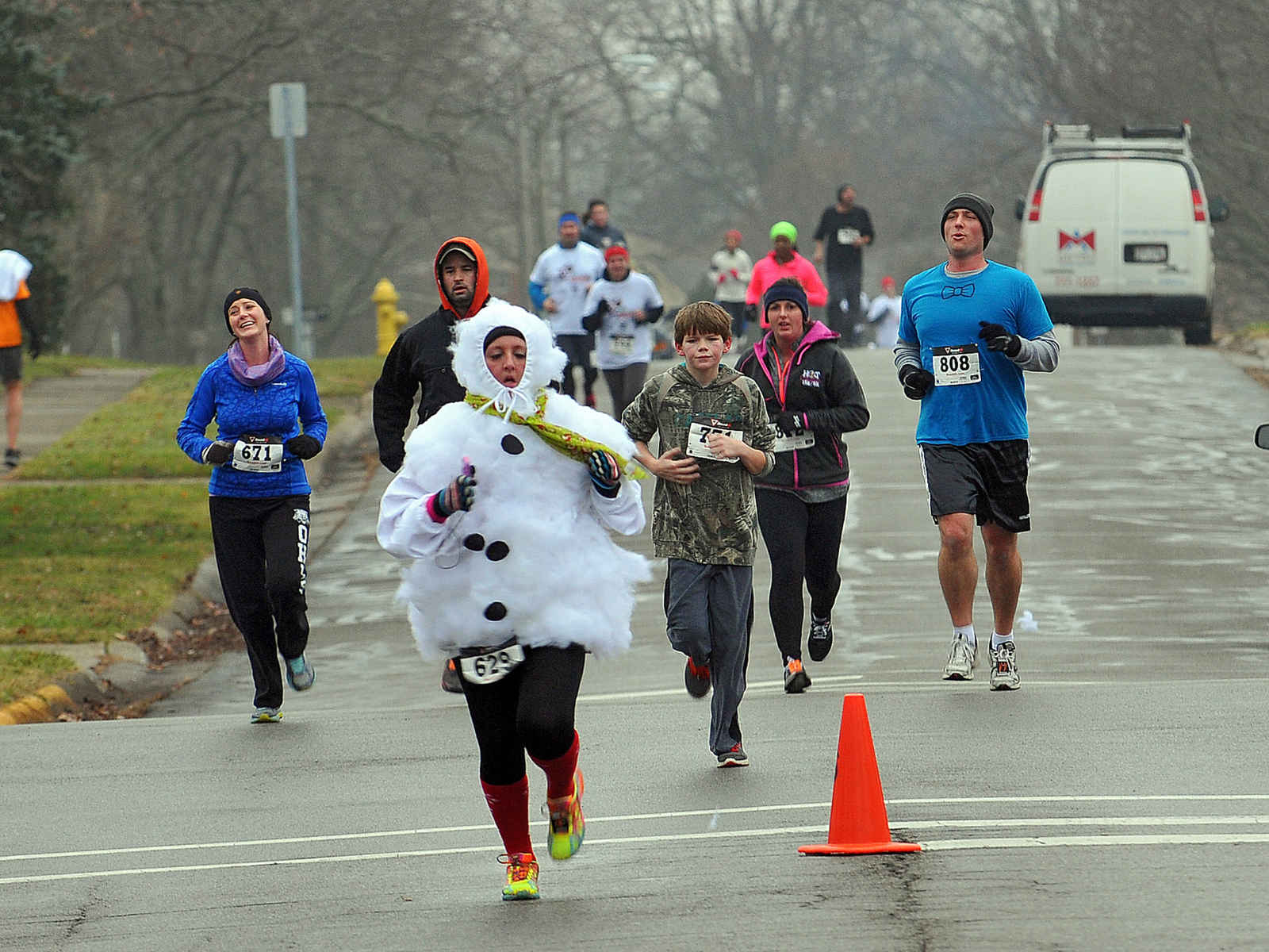 A group of runners participates in a road race on a cloudy day. One runner is wearing a snowman costume, adding a festive touch to the event. Other participants wear typical running attire. An orange traffic cone marks part of the route.
