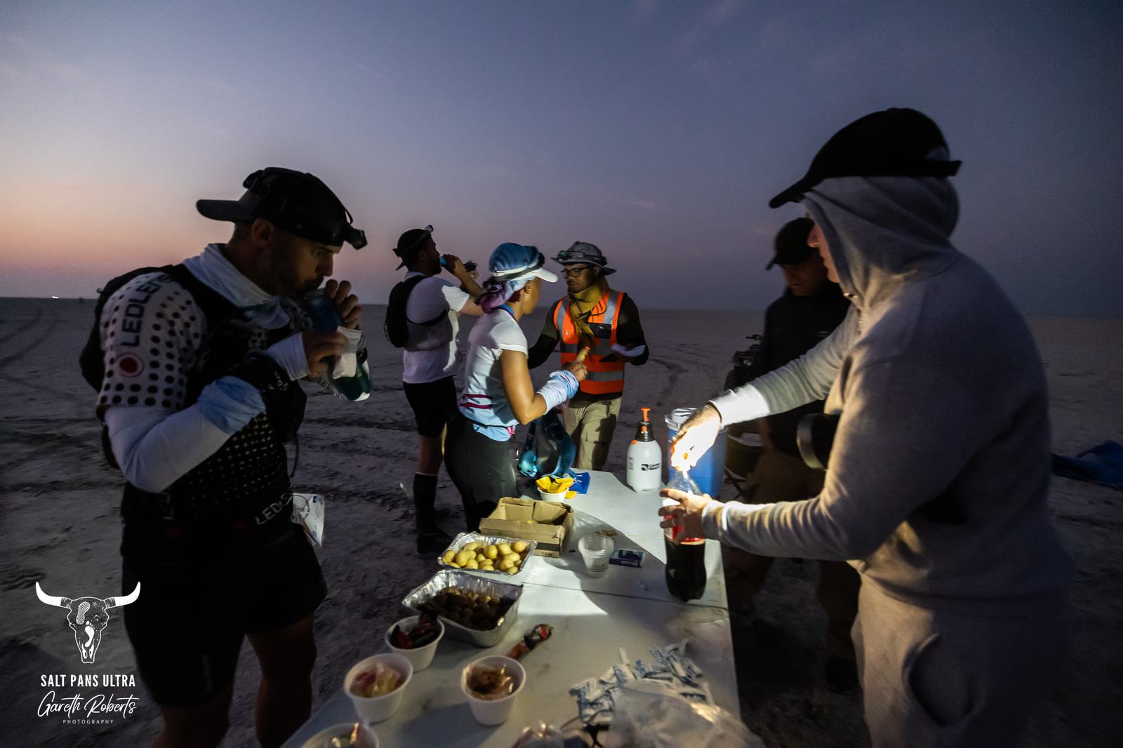 A group of people in athletic gear gather around a table with snacks and drinks in a dimly lit outdoor setting. One person pours a drink while others chat and prepare. The atmosphere appears calm and focused.