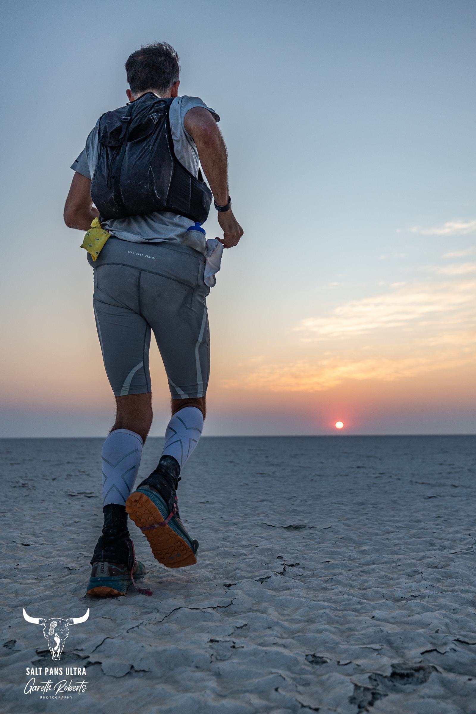 A man running on a vast, flat salt pan at sunset. He is wearing athletic gear, with a backpack and running shoes. The sky is clear with a hint of orange near the horizon where the sun is setting. Logo in the bottom corner reads "Salt Pans Ultra.