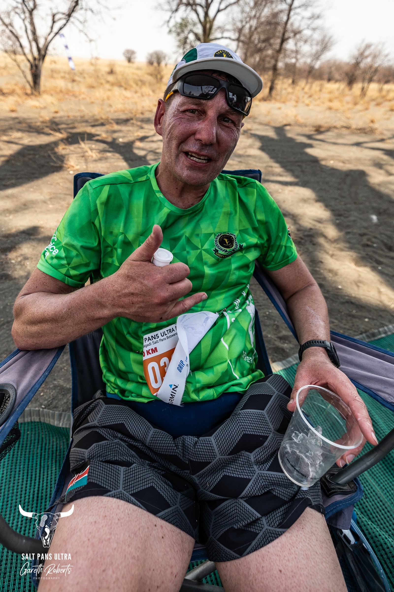 A man in a green sports shirt and patterned shorts sits in a camping chair, giving a thumbs-up. He wears sunglasses and a cap, holding a bottled drink and an empty plastic cup. Dry landscape with trees in the background.