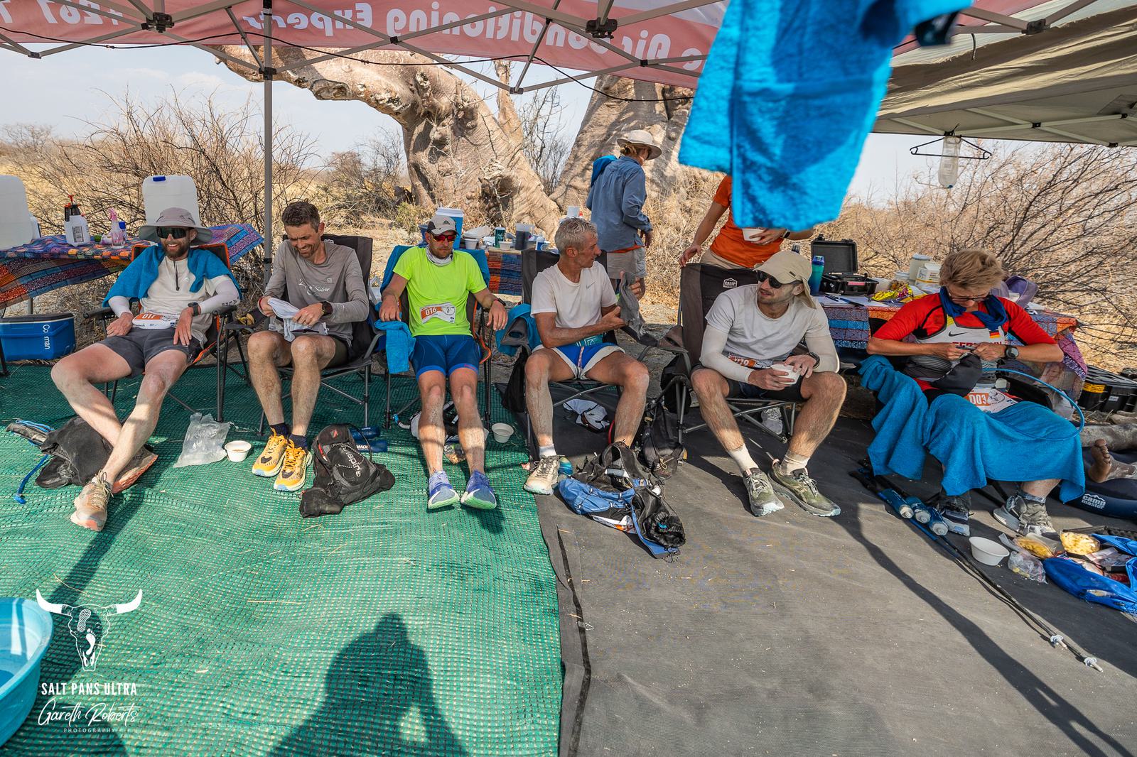 A group of six people sit on folding chairs under a canopy in a dry outdoor setting. They appear to be resting, with some looking down and others reading. A large tree is visible in the background. Various supplies and equipment are scattered around.