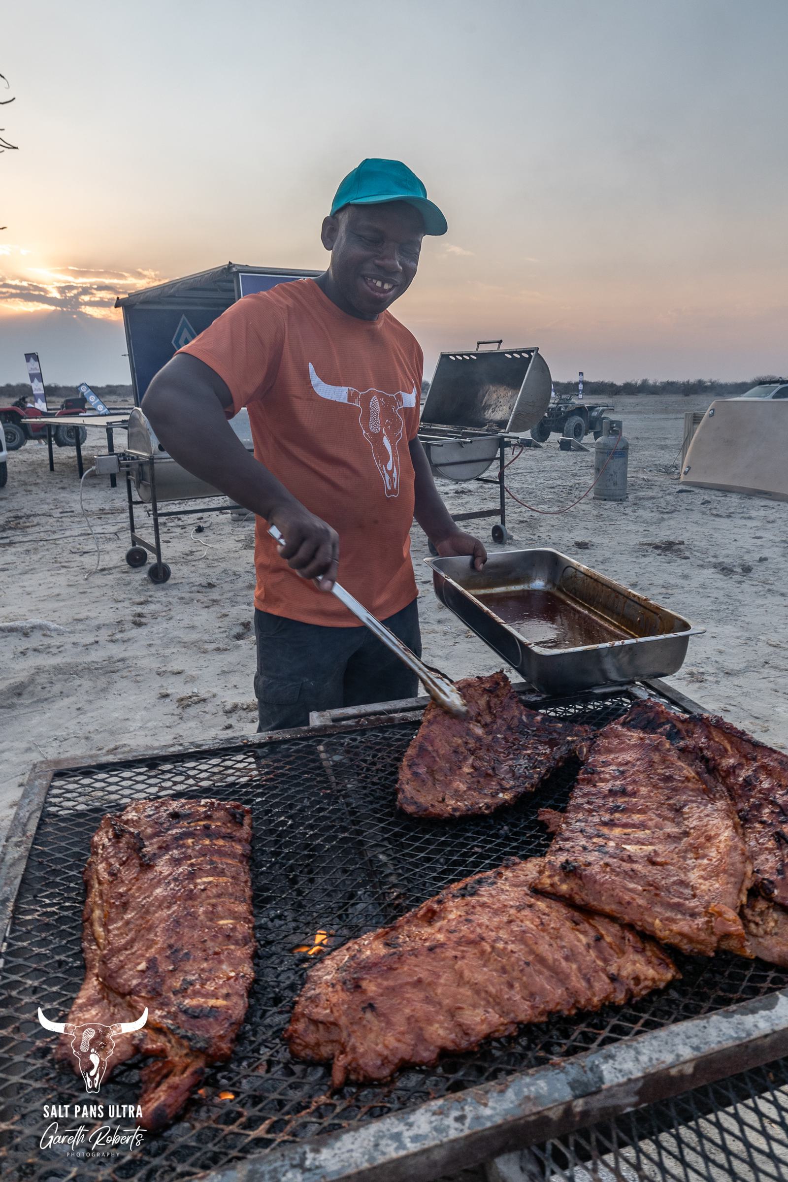 A man in an orange shirt and green cap grills racks of ribs on an outdoor barbecue. He smiles while holding a pan beside the grill. In the background, there are vehicles and cooking equipment on a sandy terrain under a cloudy sky.