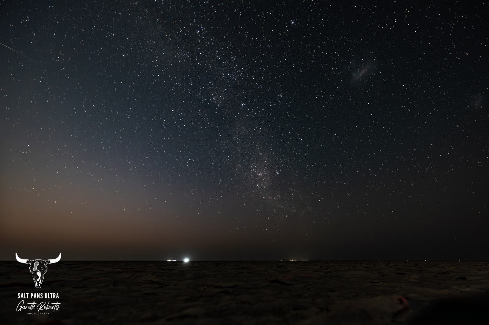 A vast night sky filled with countless stars twinkles over a dark horizon. Dim lights appear on the horizon, while a faint glow illuminates part of the sand below. A branded logo is visible in the bottom left corner.