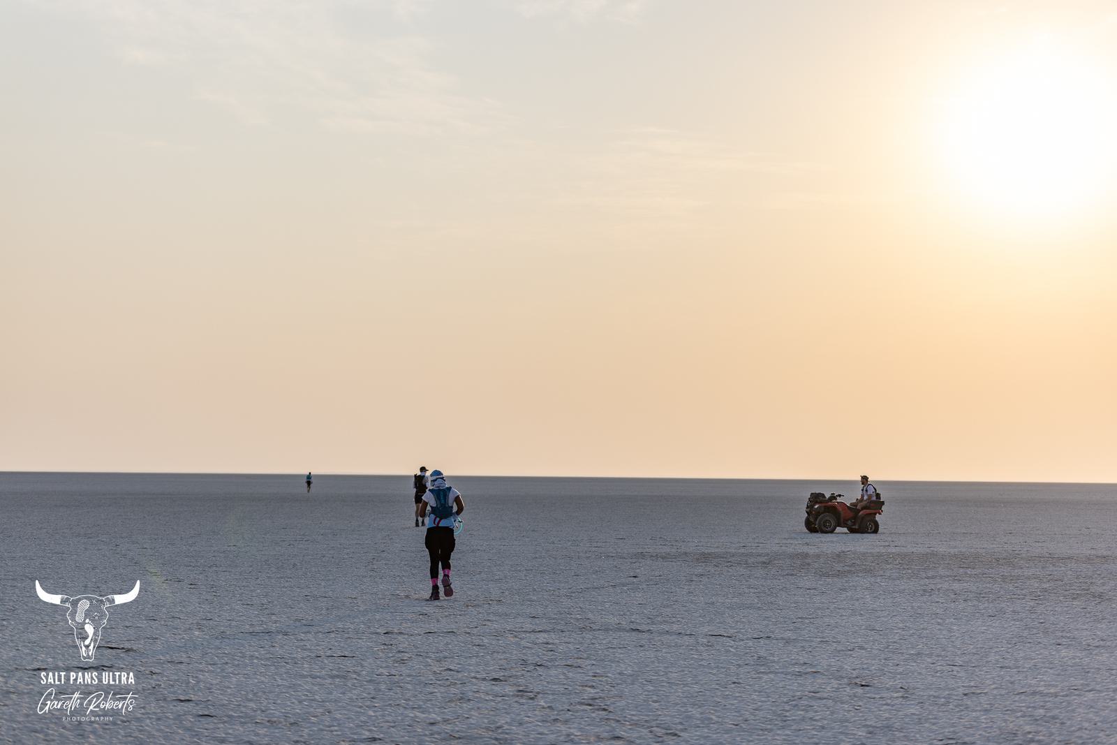 A person in athletic gear runs across a vast, flat, and barren landscape at sunrise. In the distance, a vehicle is parked with another figure nearby. The sky is clear with a few clouds near the horizon.